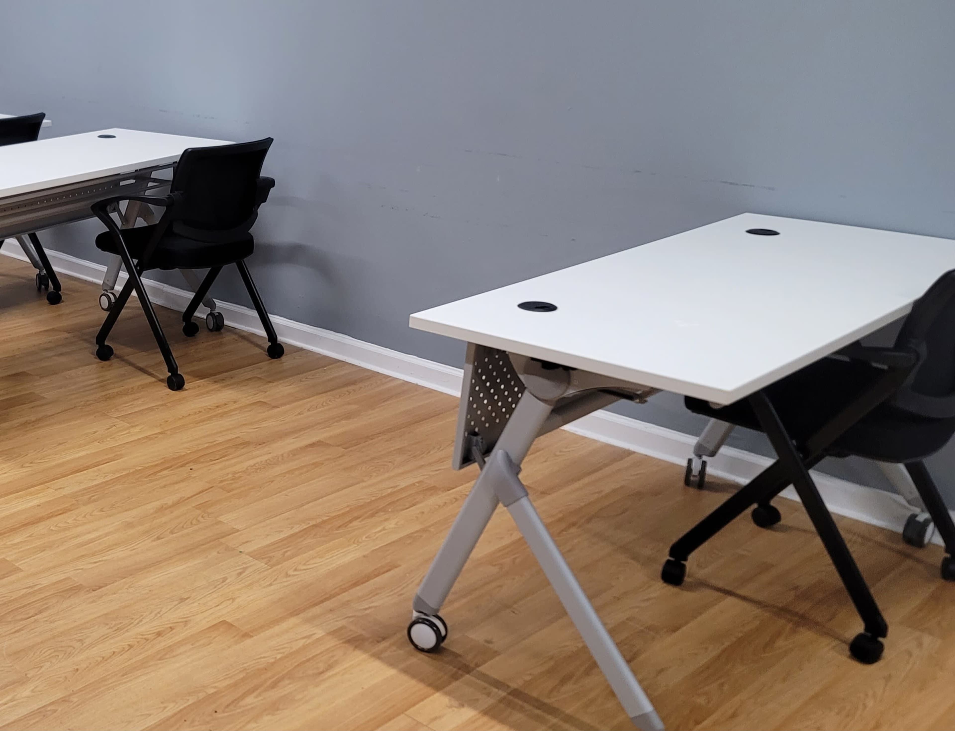 A workspace with multiple white tables and rolling black chairs on a wooden floor, positioned against a gray wall.