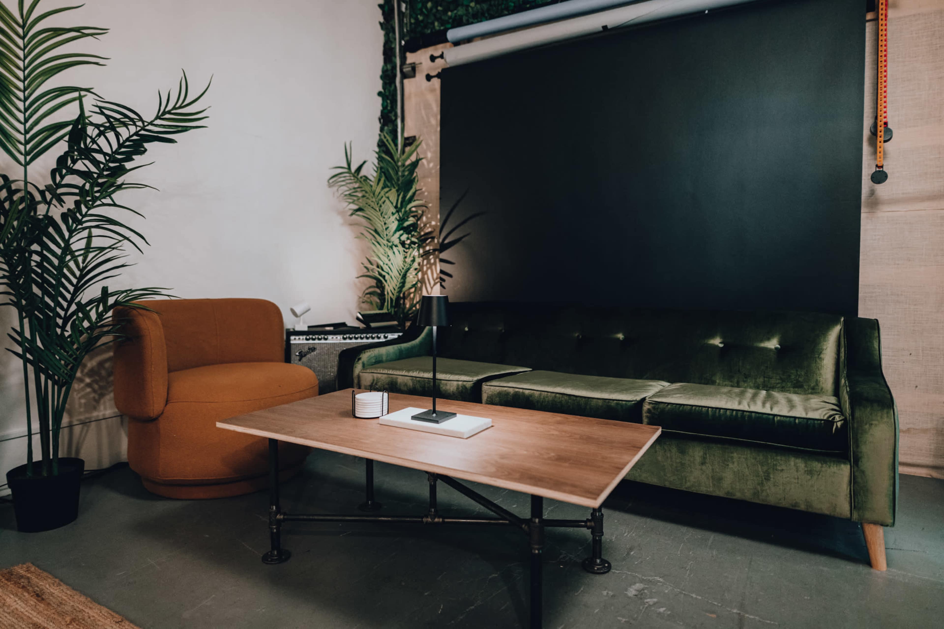 A green velvet sofa and a round orange chair are positioned around a wooden coffee table in a room with potted plants and a black backdrop.