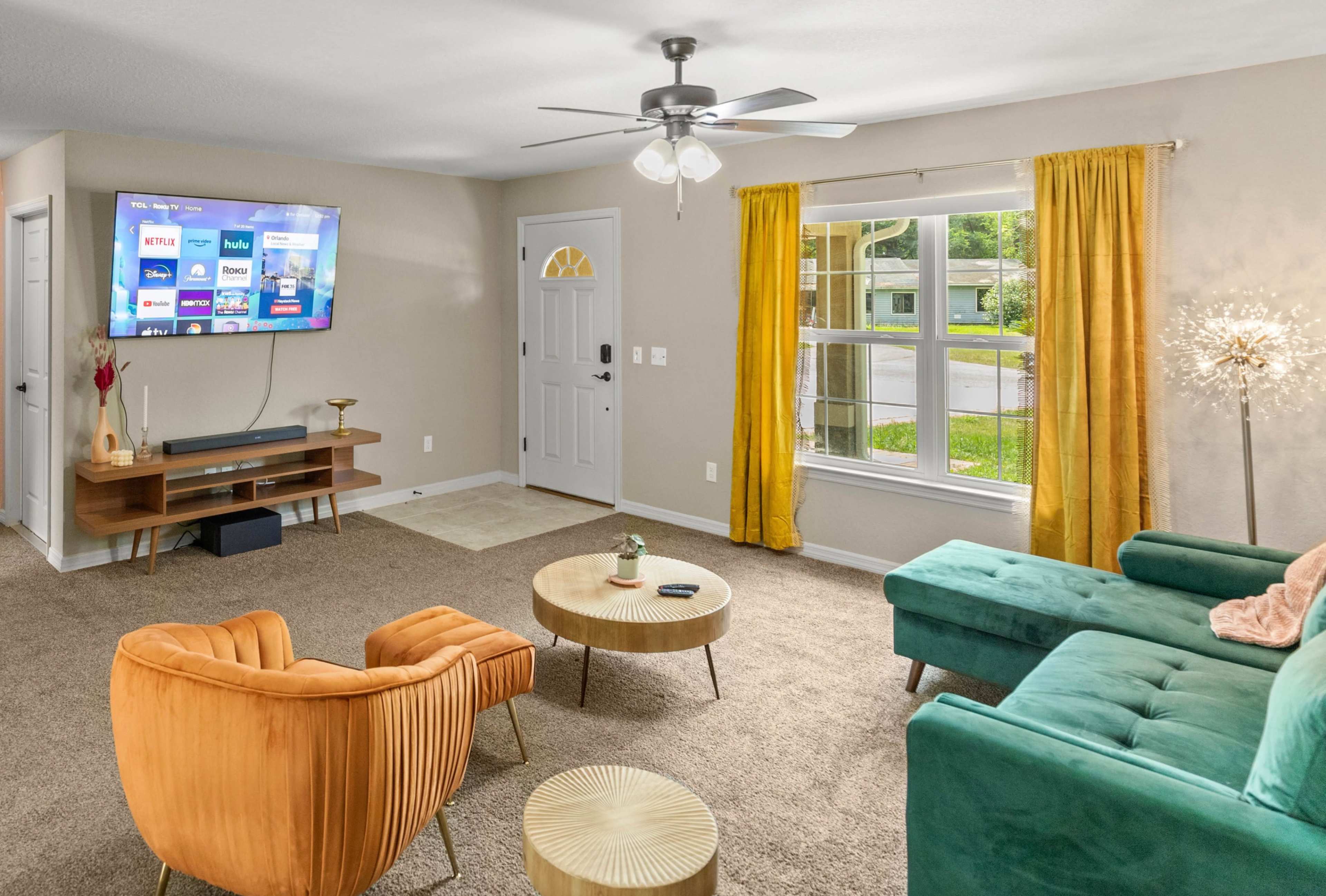 The living room features a television mounted on the wall, a green sofa, two orange chairs, and a large window with yellow curtains.