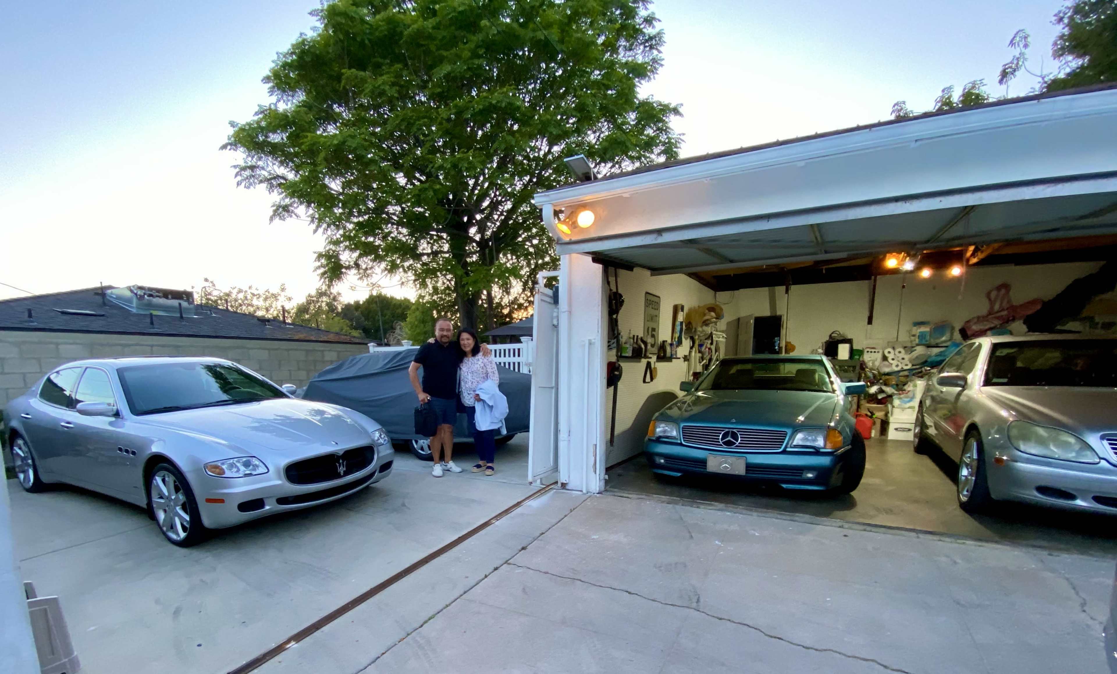 A couple stands in front of a garage beside two parked cars, one a silver Maserati and the other a blue Mercedes-Benz, with additional vehicles visible inside the garage.