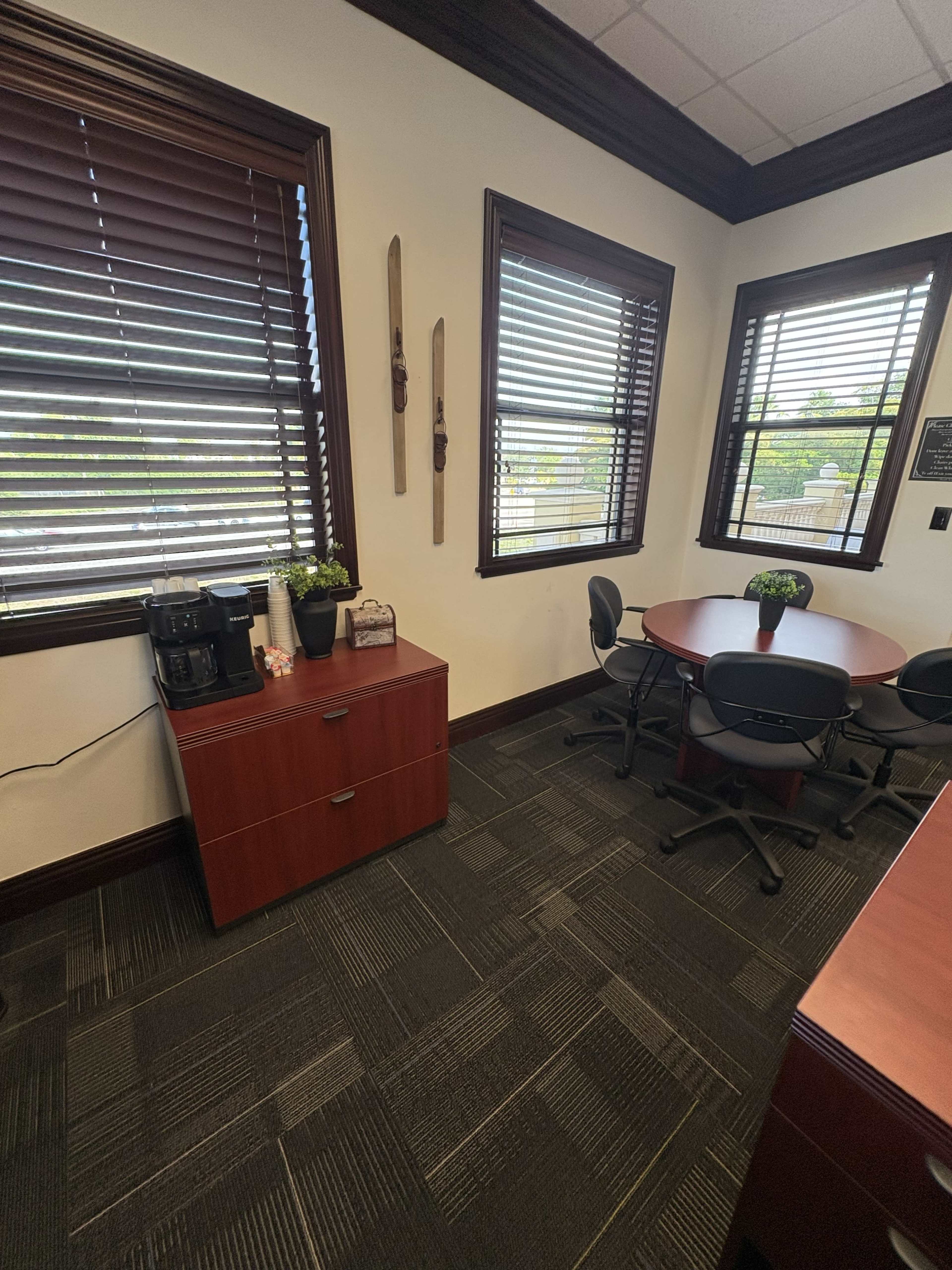 The image shows a small office with a coffee machine on a cabinet, a round table with chairs, and wooden blinds on the windows.