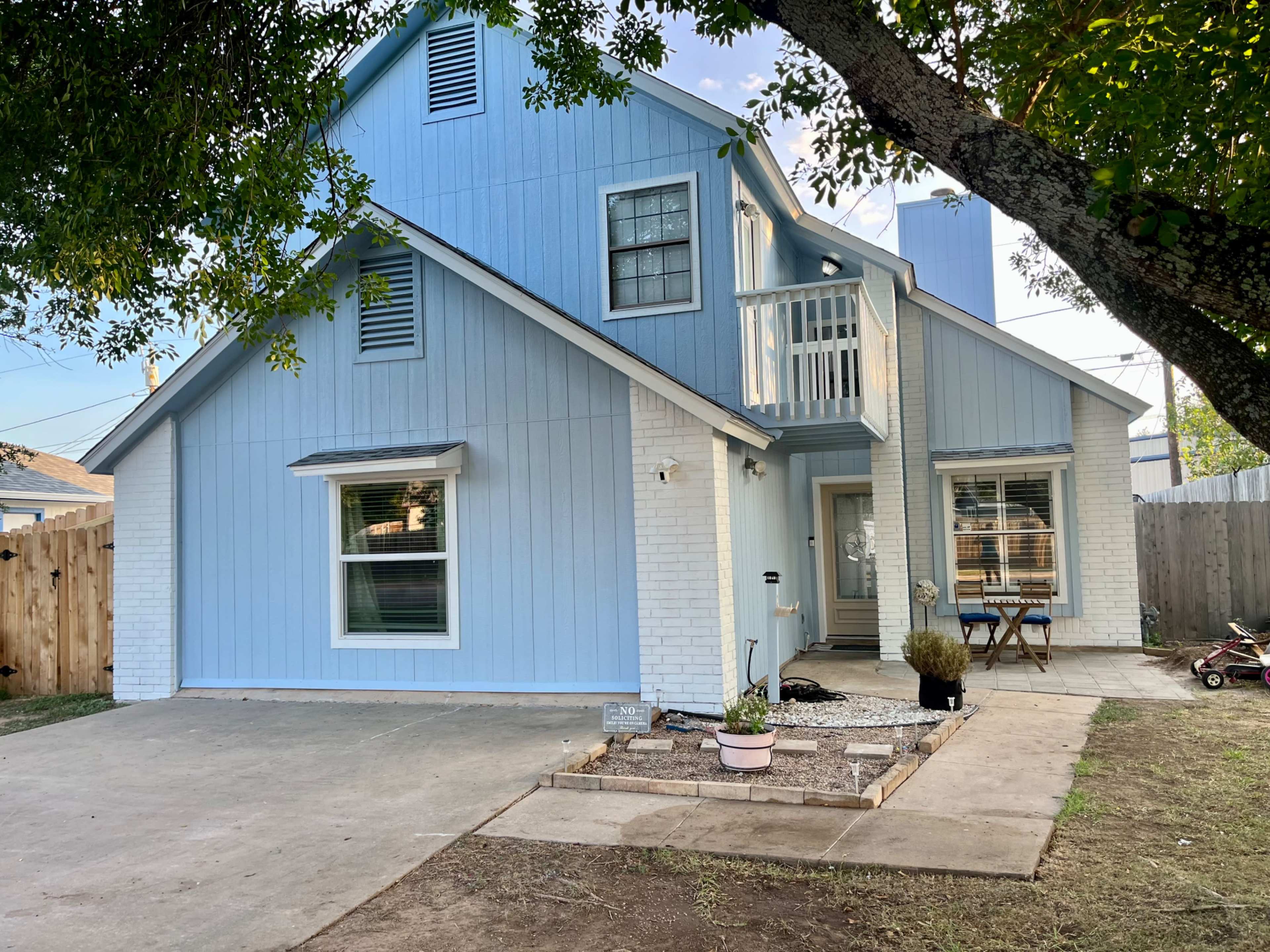 A two-story blue house with white trim features a front porch and a small garden area, situated on a concrete driveway.