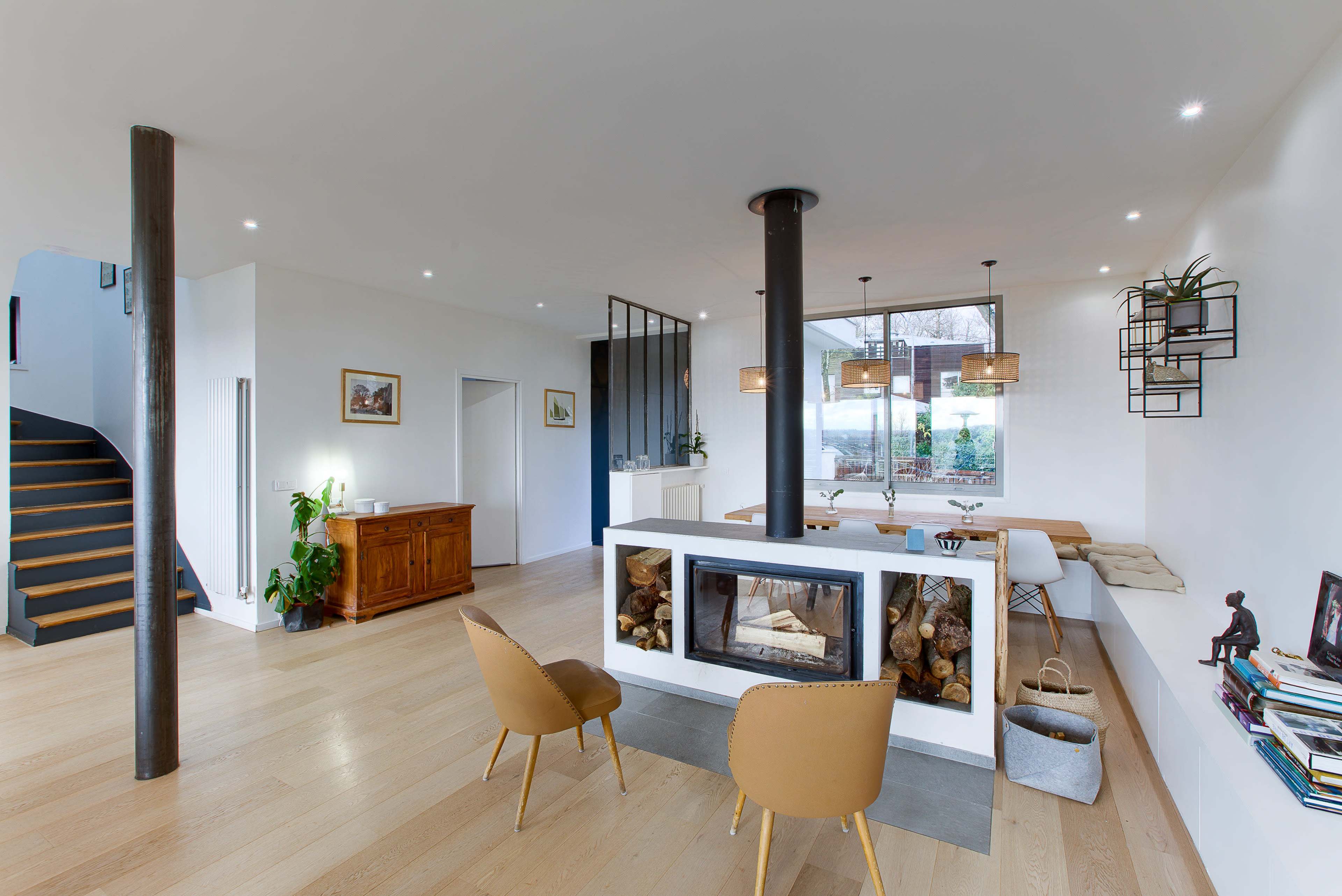 The image shows a modern open-concept living space that features a central fireplace, a dining area with a large window, and a staircase in the background.