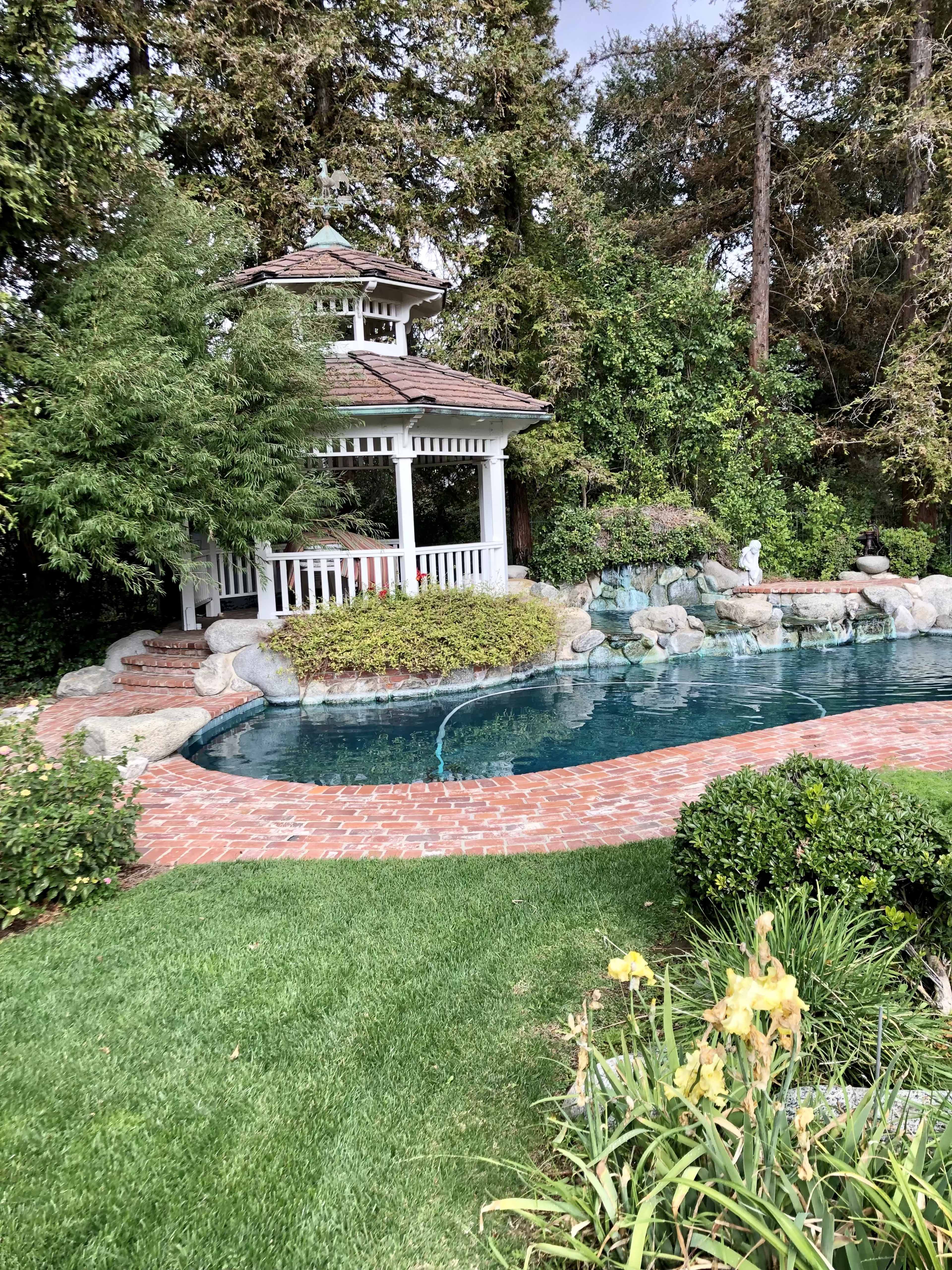 The image shows a landscaped backyard featuring a swimming pool bordered by brick pathways and a gazebo surrounded by lush greenery.