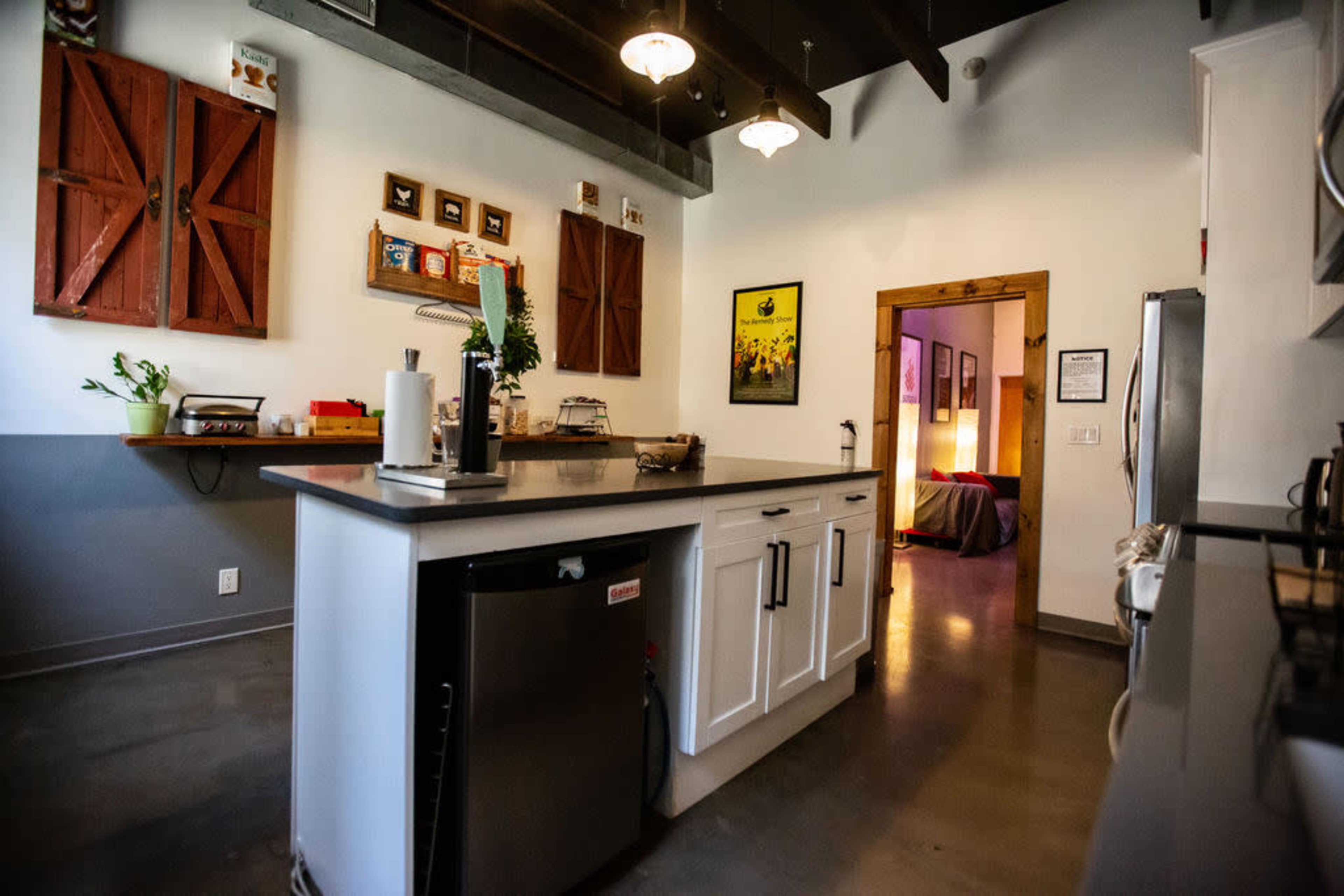 The image shows a modern kitchen with a central island, featuring a stainless steel refrigerator and rustic wooden shutters on the walls, leading to a softly lit hallway with a bedroom in the background.