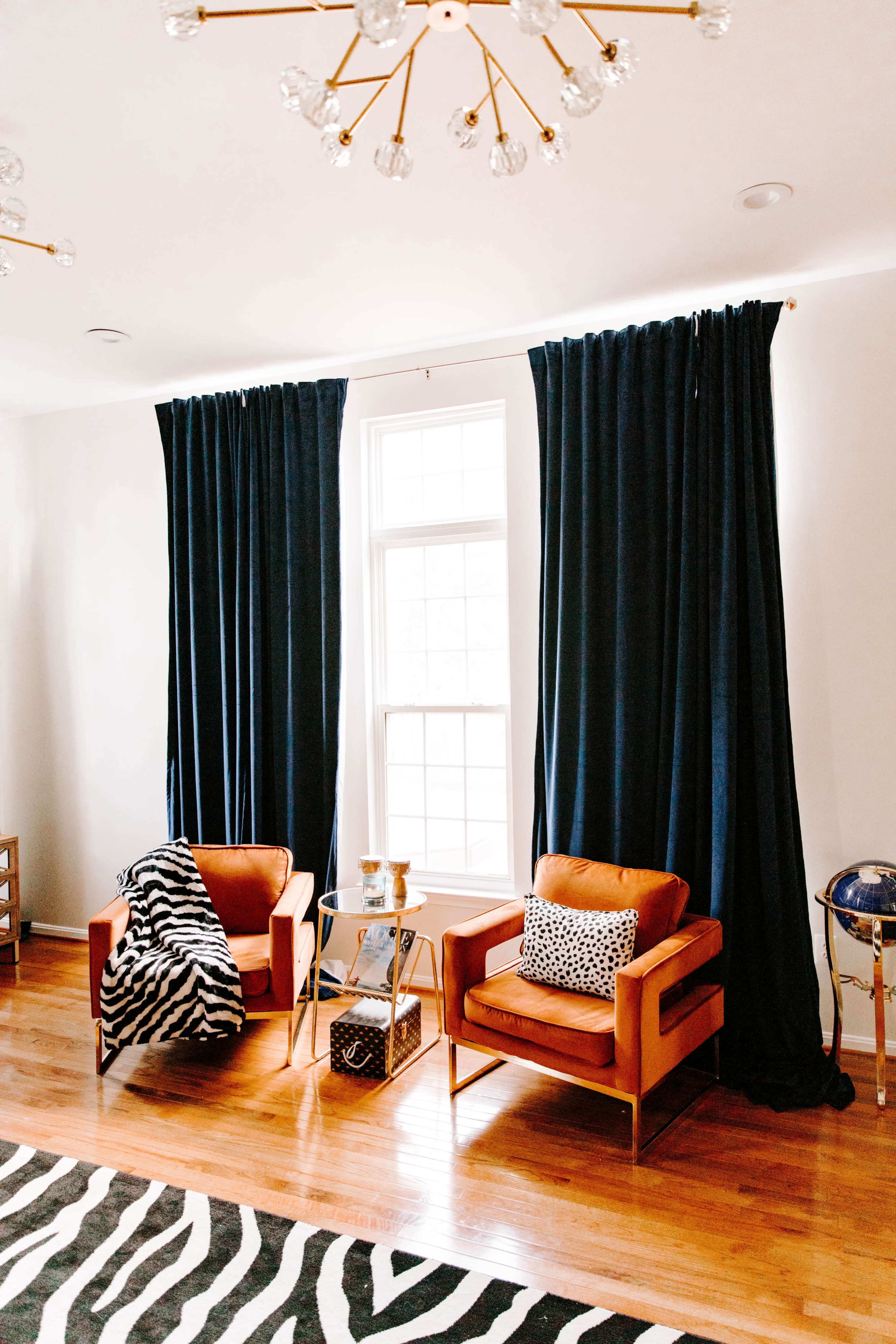 A living room with two orange chairs, a small glass side table, and dark blue curtains hanging over a window, set against a zebra-striped rug on a wooden floor.