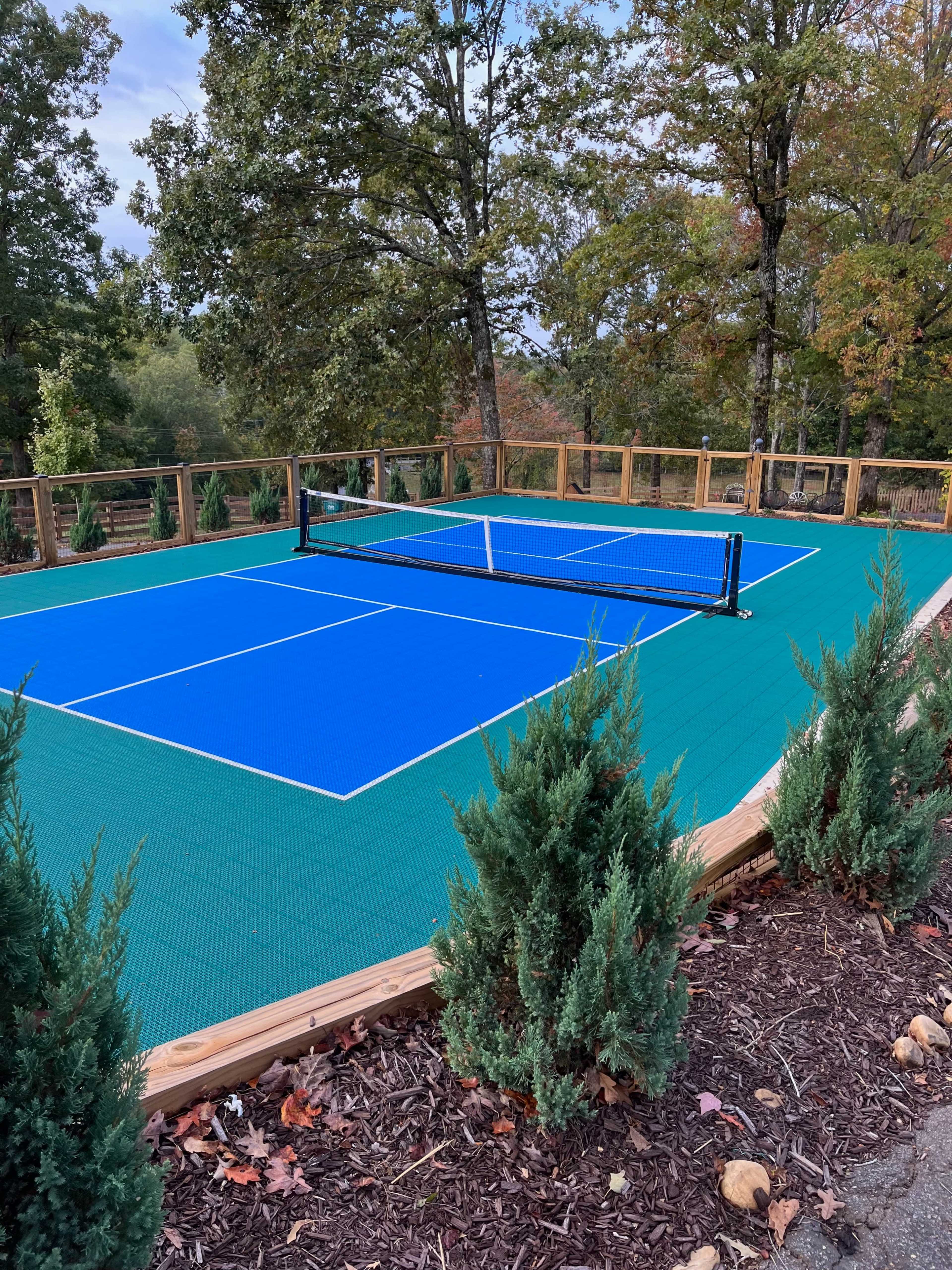 A blue tennis court surrounded by greenery and wooden fencing is located in a forested area.