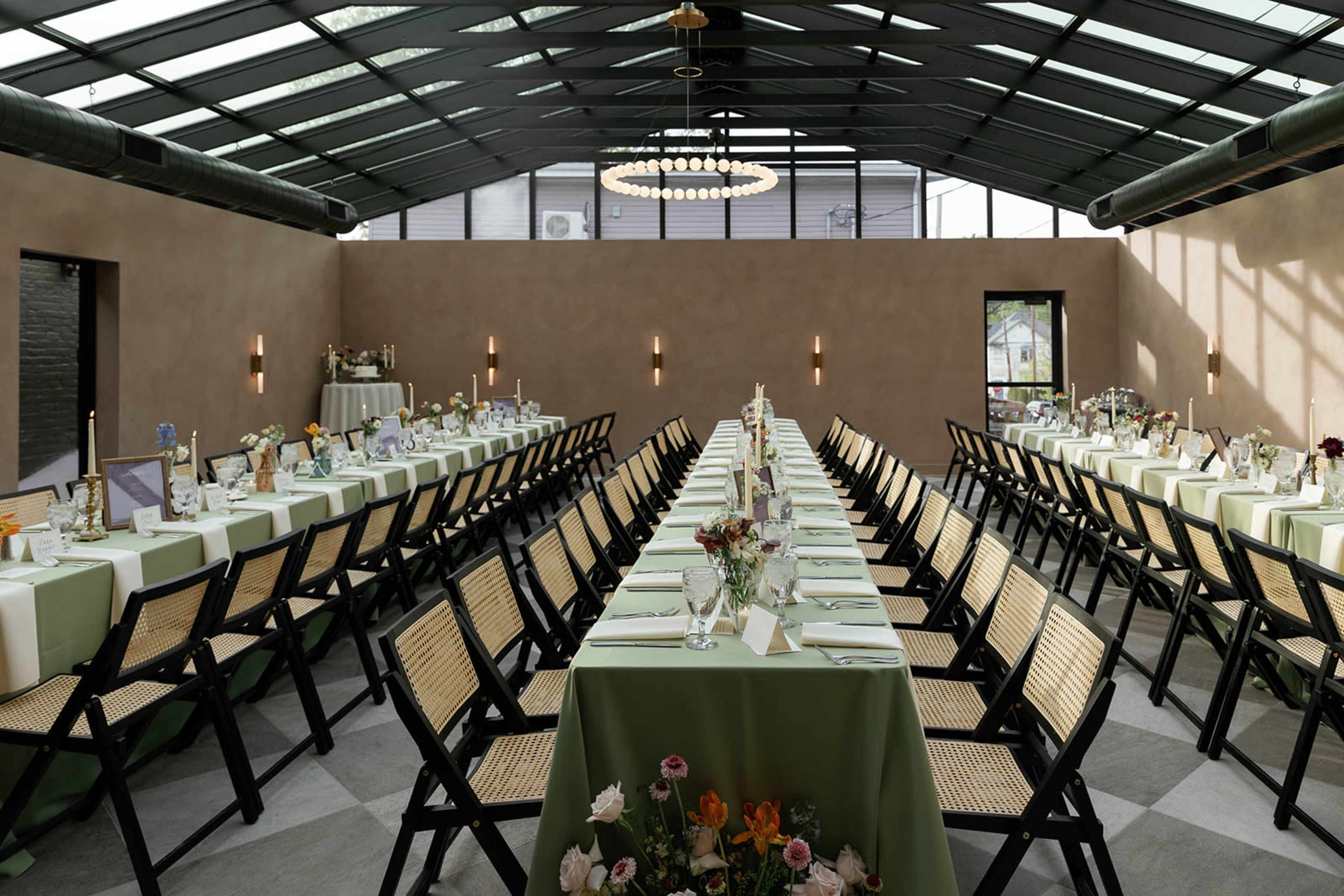 A spacious dining area set with long tables covered in green tablecloths, adorned with floral centerpieces and lit by an overhead chandelier.