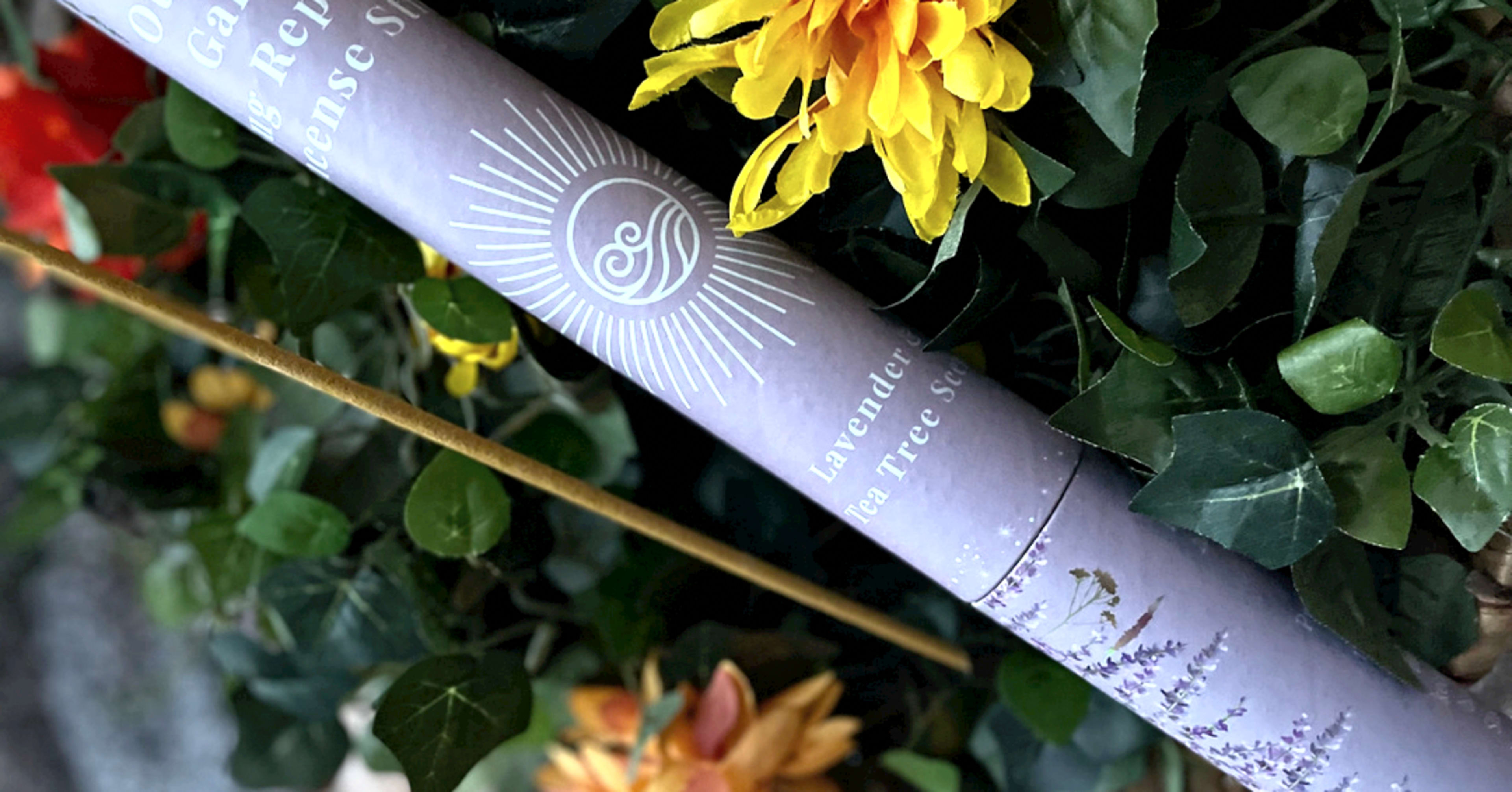 A lavender and tea tree scented incense stick is partially visible among vibrant flowers and green leaves.