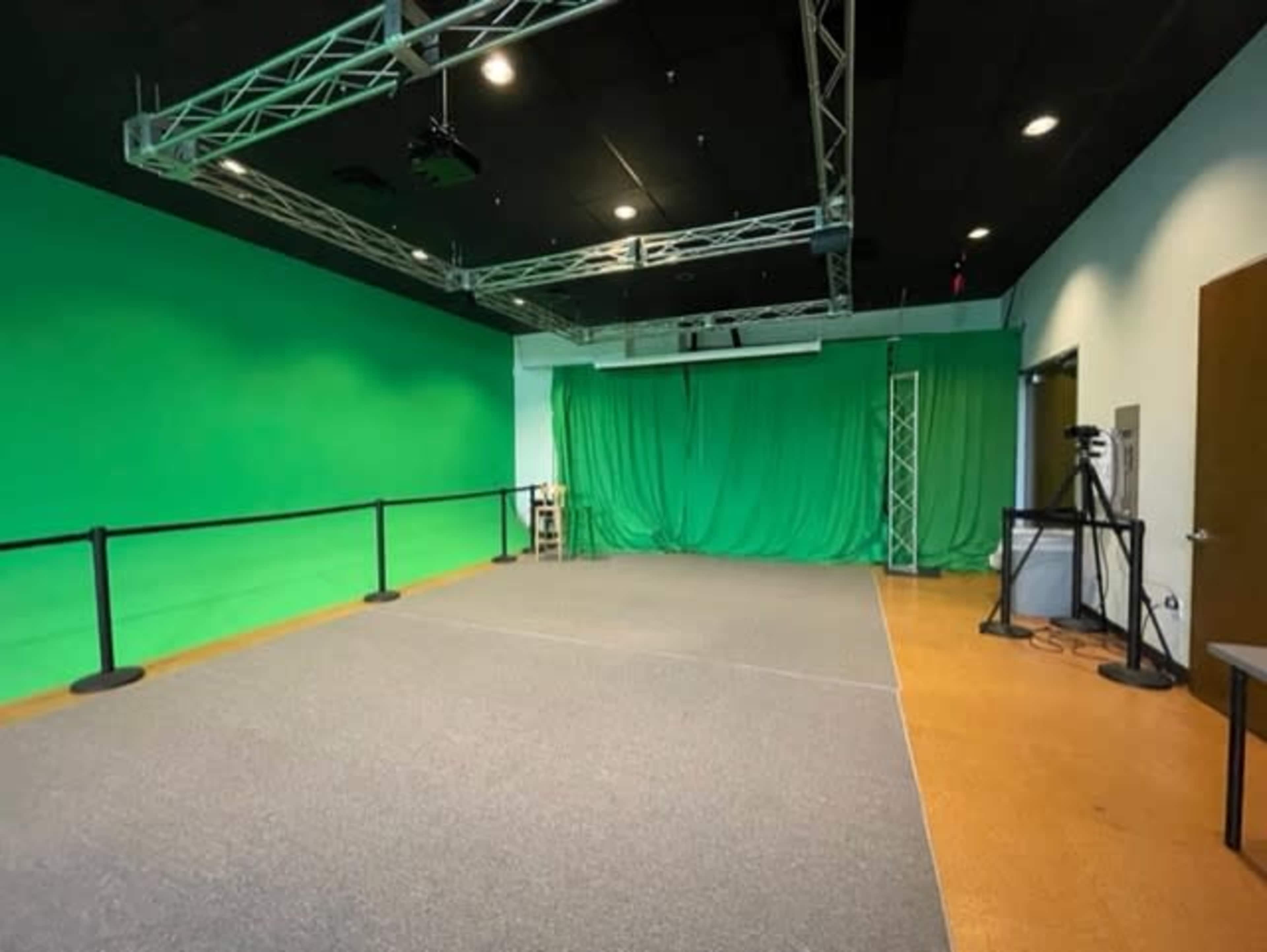 A spacious green screen studio with a gray carpet and lighting rig set up in the ceiling.