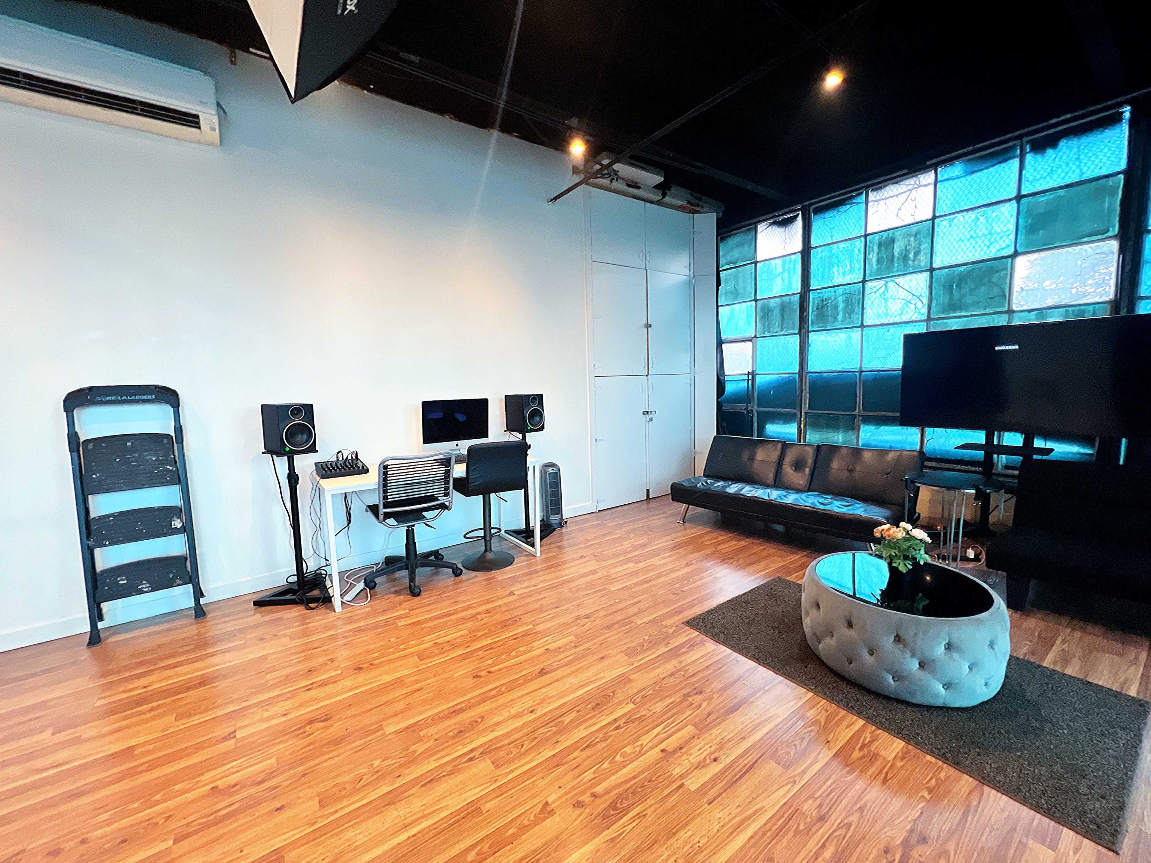 The image shows a modern studio space with a black sofa, a round coffee table, and audio equipment against a backdrop of large windows.