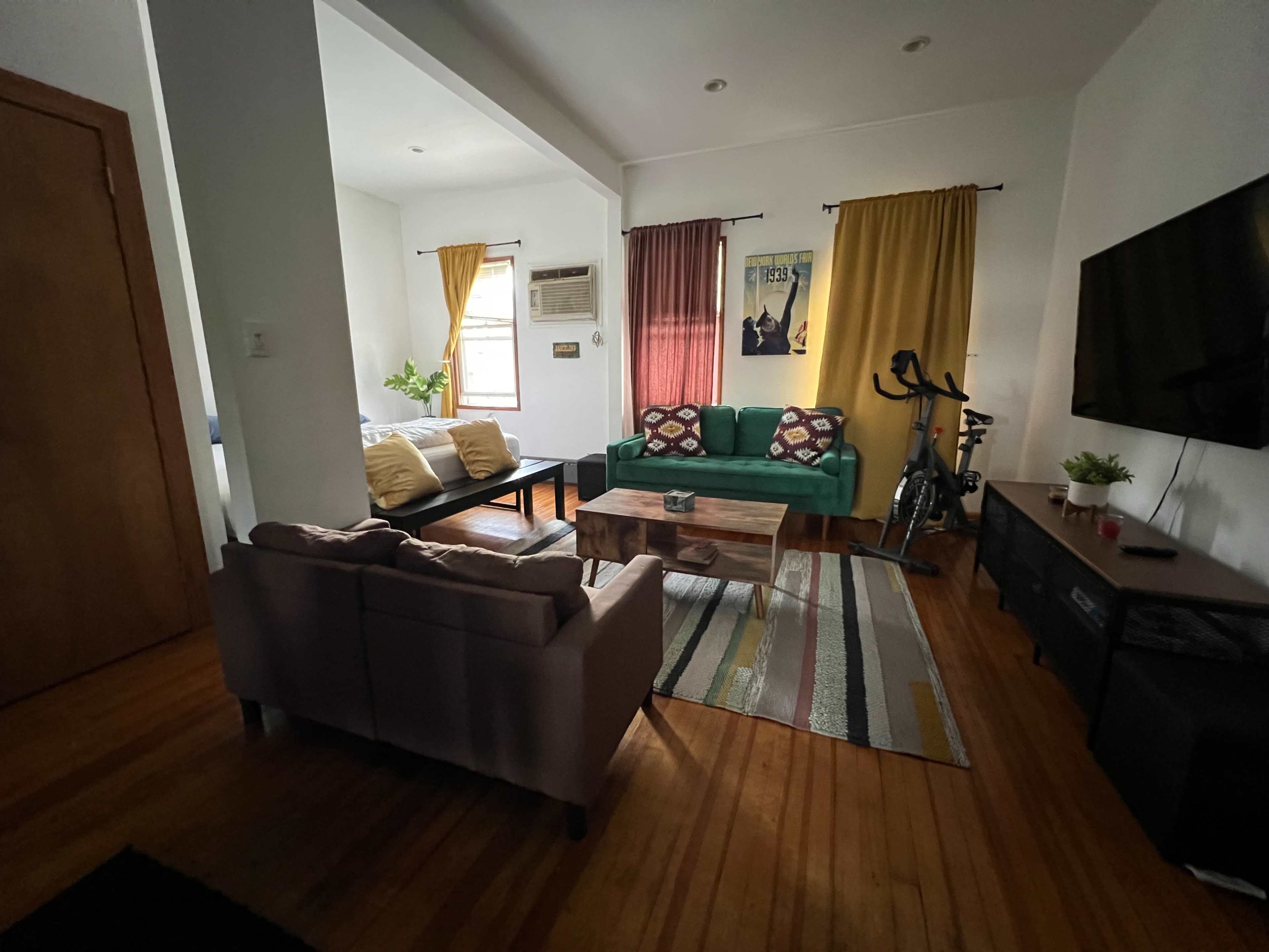 The image shows a living room with a green couch, a coffee table, and a bike beside a TV unit, featuring window curtains and wooden flooring.