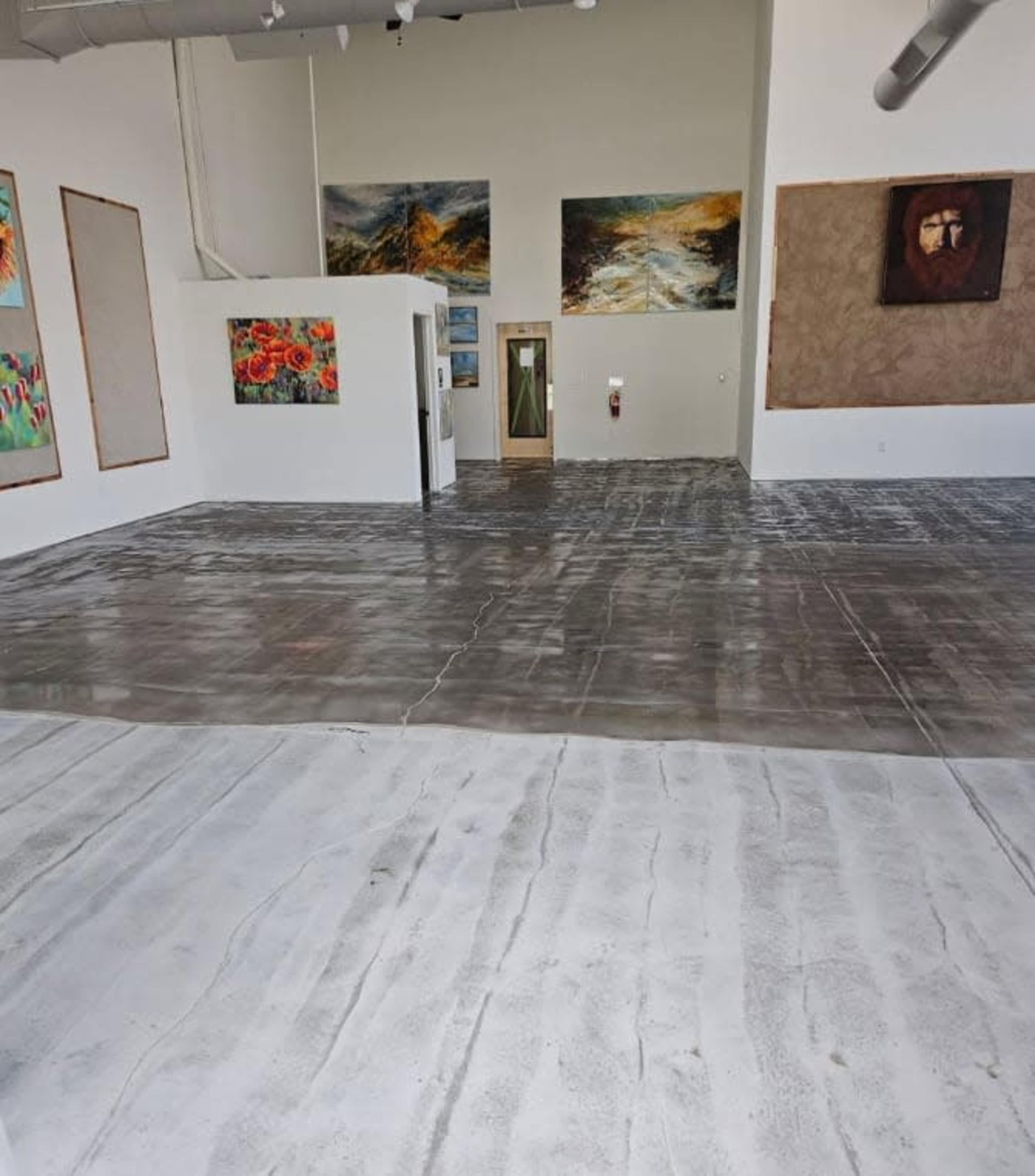 The image shows an empty art gallery with concrete floors and various artworks displayed on the walls.