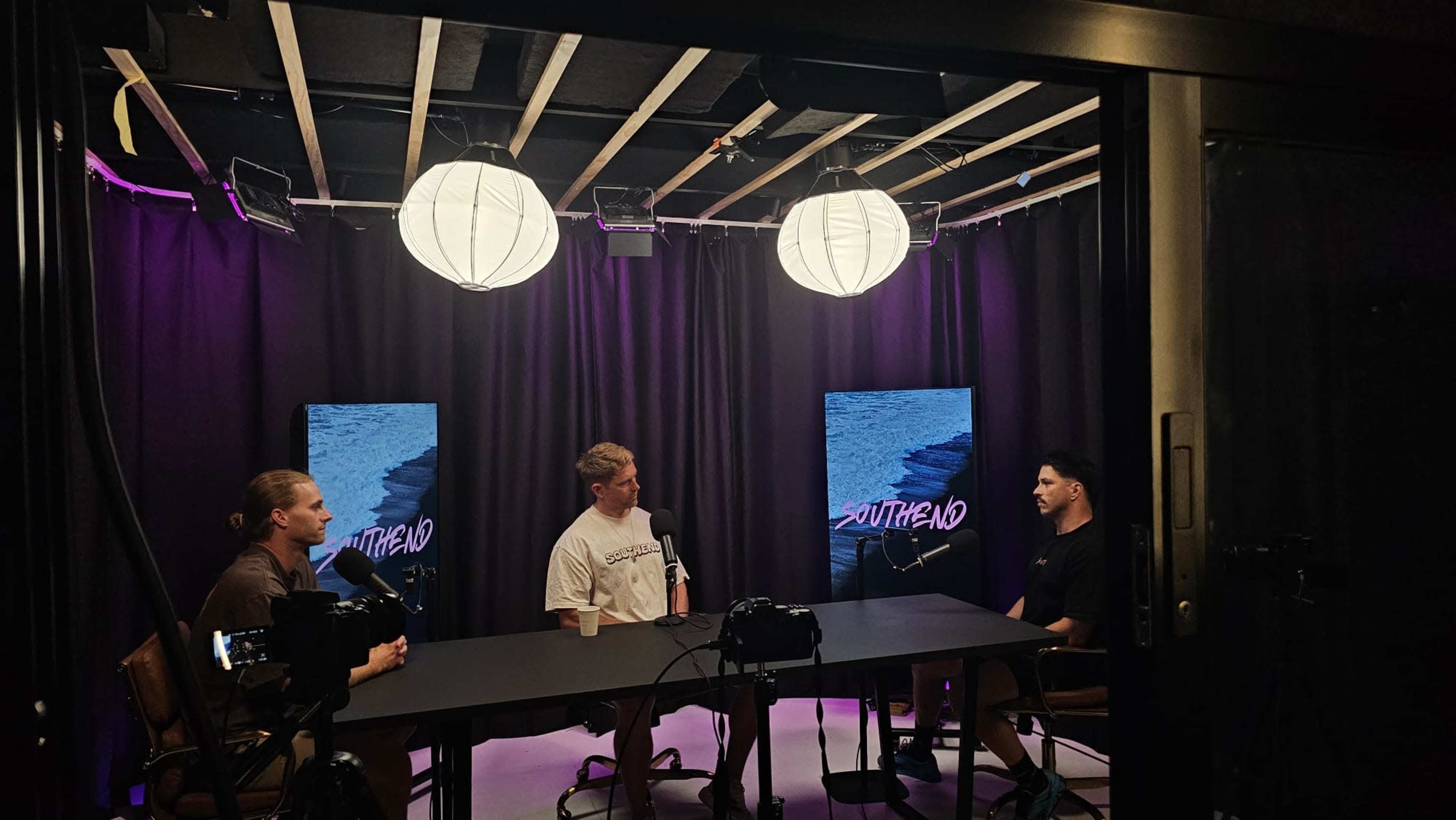 Three men sit at a table in a recording studio, surrounded by black curtains and lit by hanging paper lanterns, with two screens displaying the word "SOUTHBOUND" in the background.