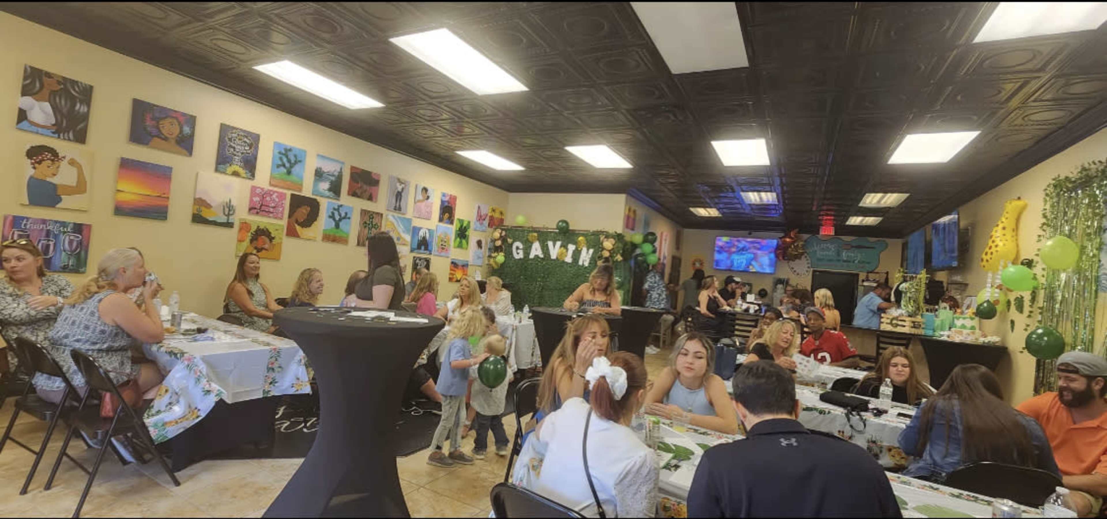 A lively indoor event with guests seated at tables decorated with festive tablecloths, surrounded by artwork on the walls and balloons.