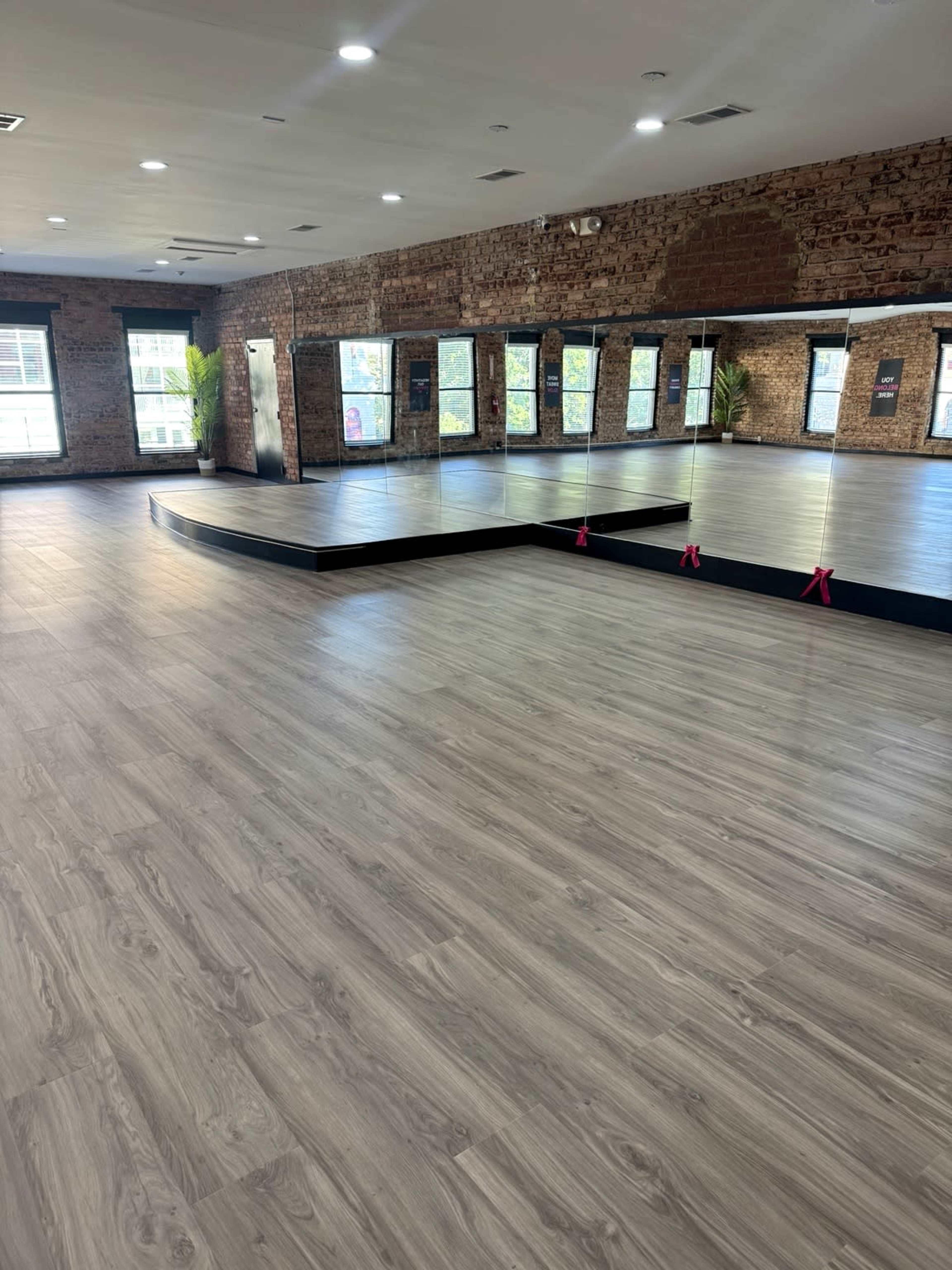 The image depicts a spacious dance studio with wooden flooring, large mirrors, and exposed brick walls.