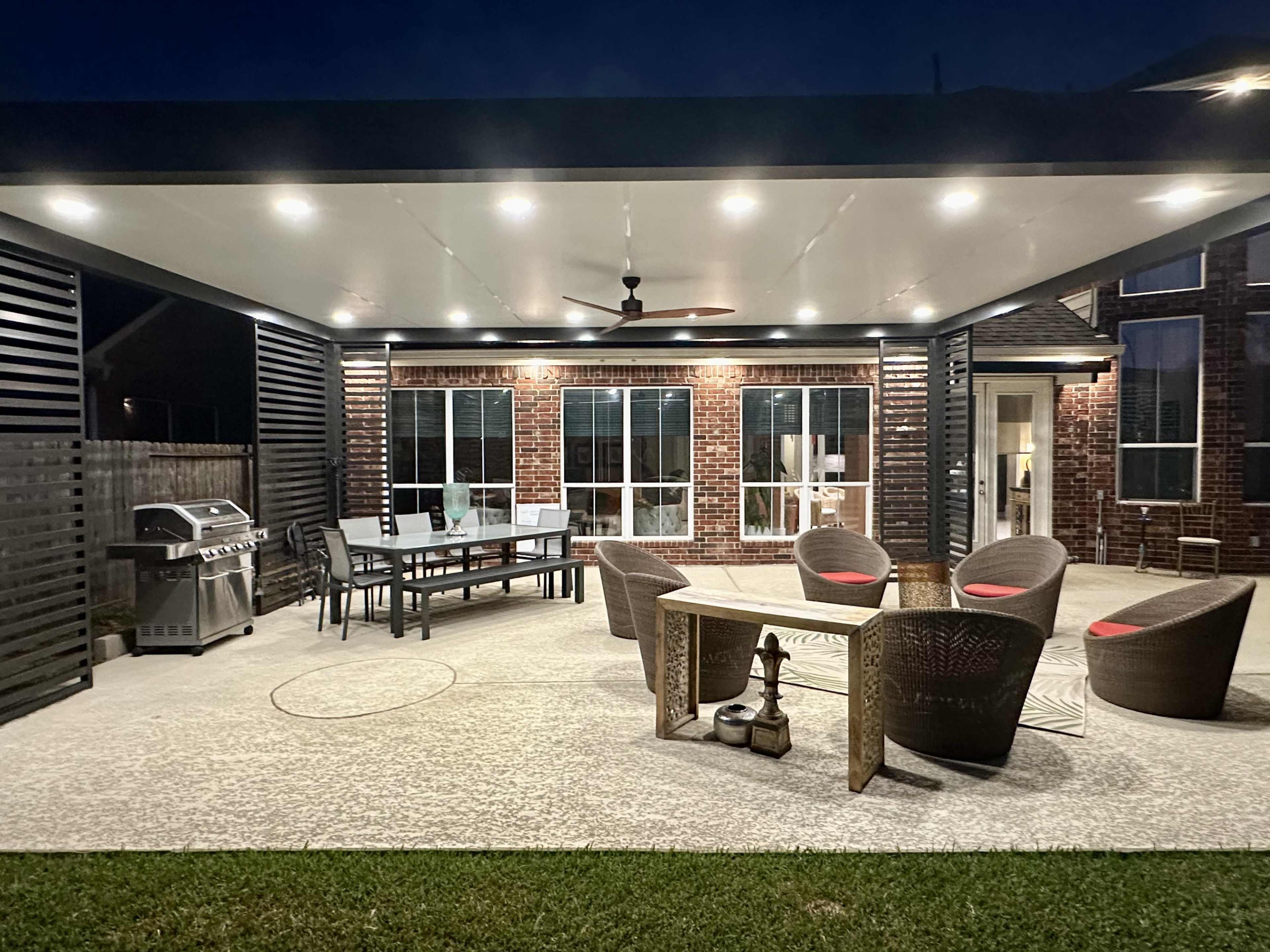 A modern outdoor patio features a dining table, grill, and seating area under a roof with overhead lights.
