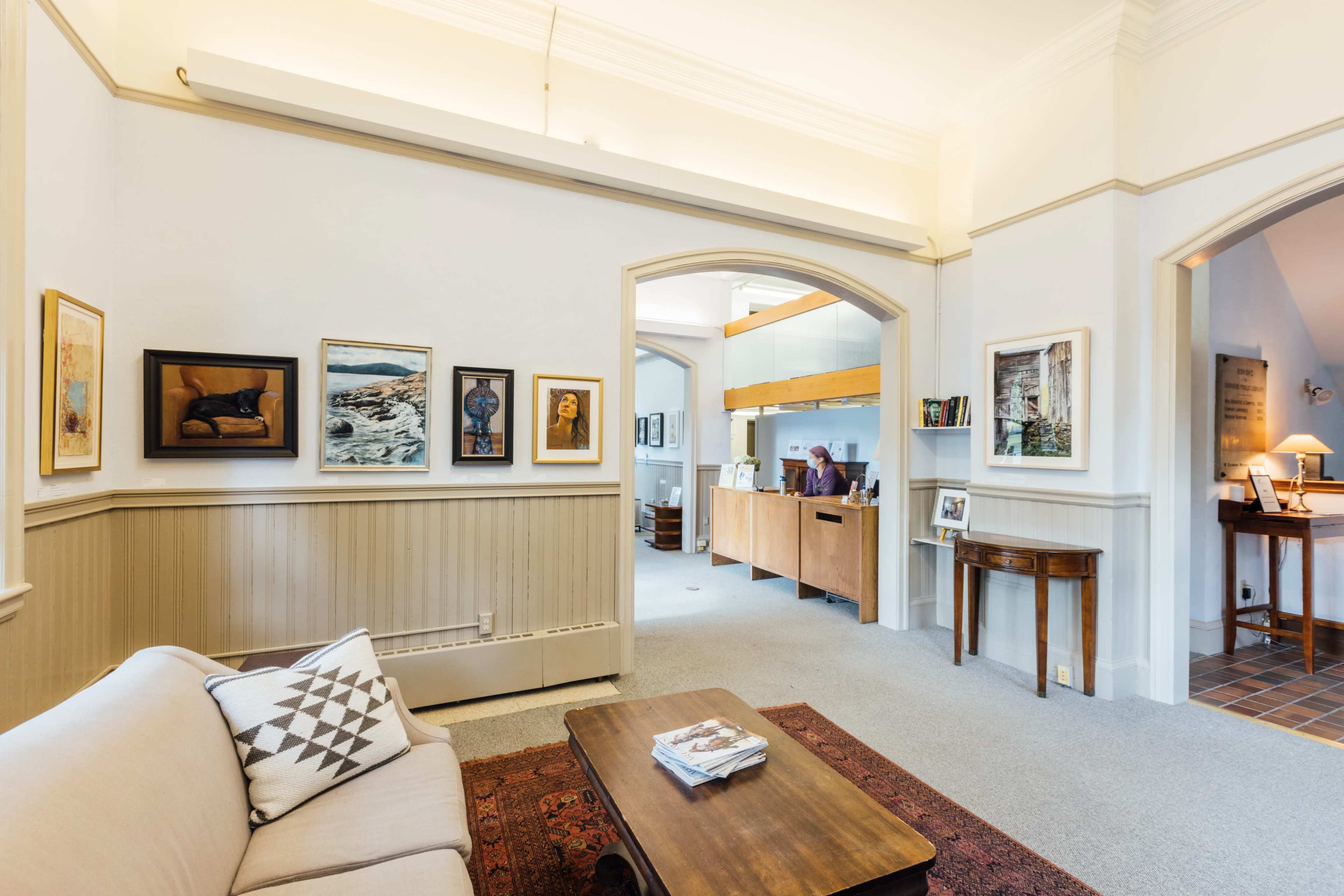 A bright room featuring a sofa, a coffee table, and walls adorned with framed artwork, leading to an adjacent space with a reception area and additional furnishings.