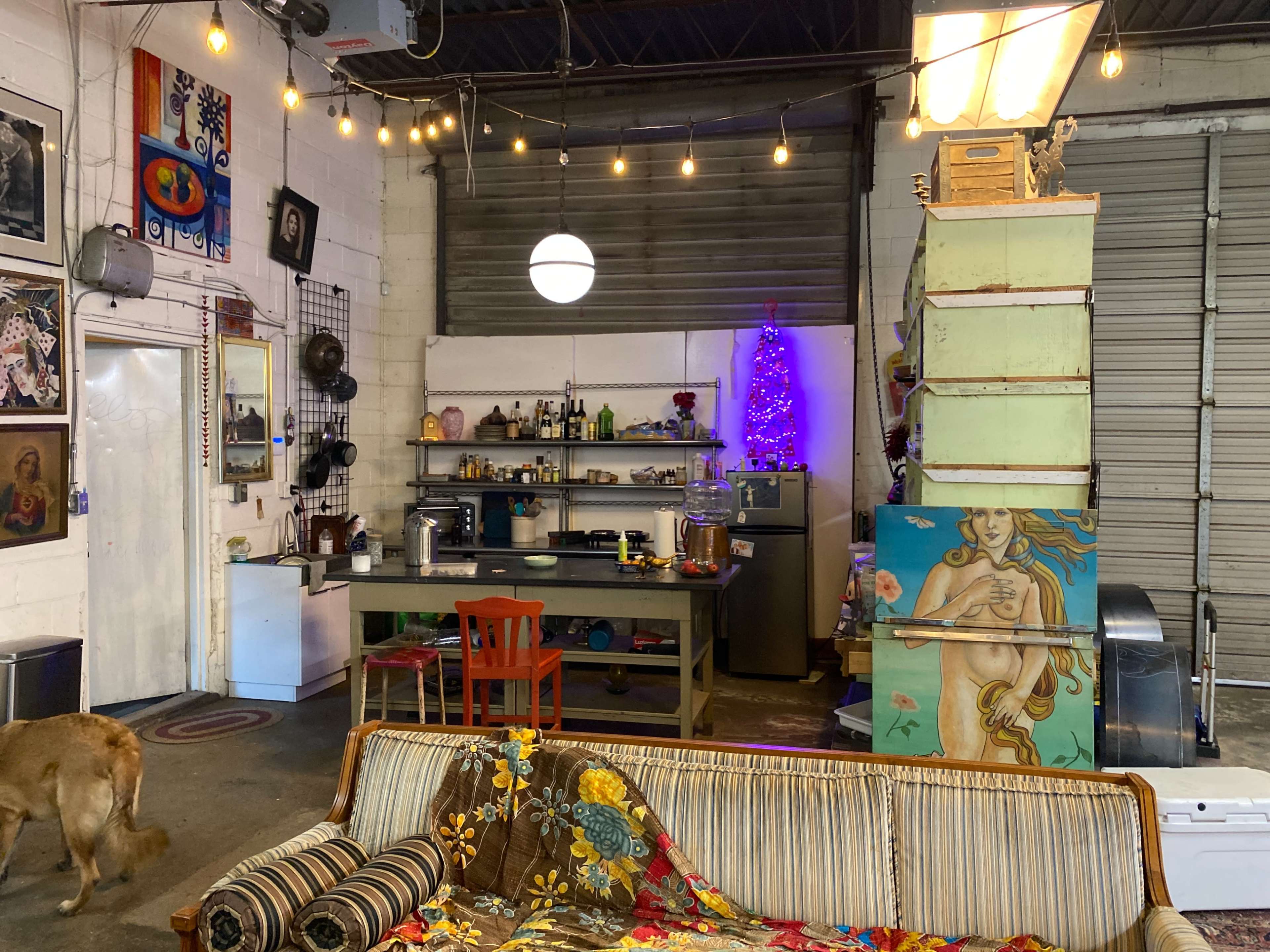 The image shows a cozy industrial-style space featuring a kitchen area with shelves, a dining table, a decorative wall, and a couch adorned with a colorful blanket.