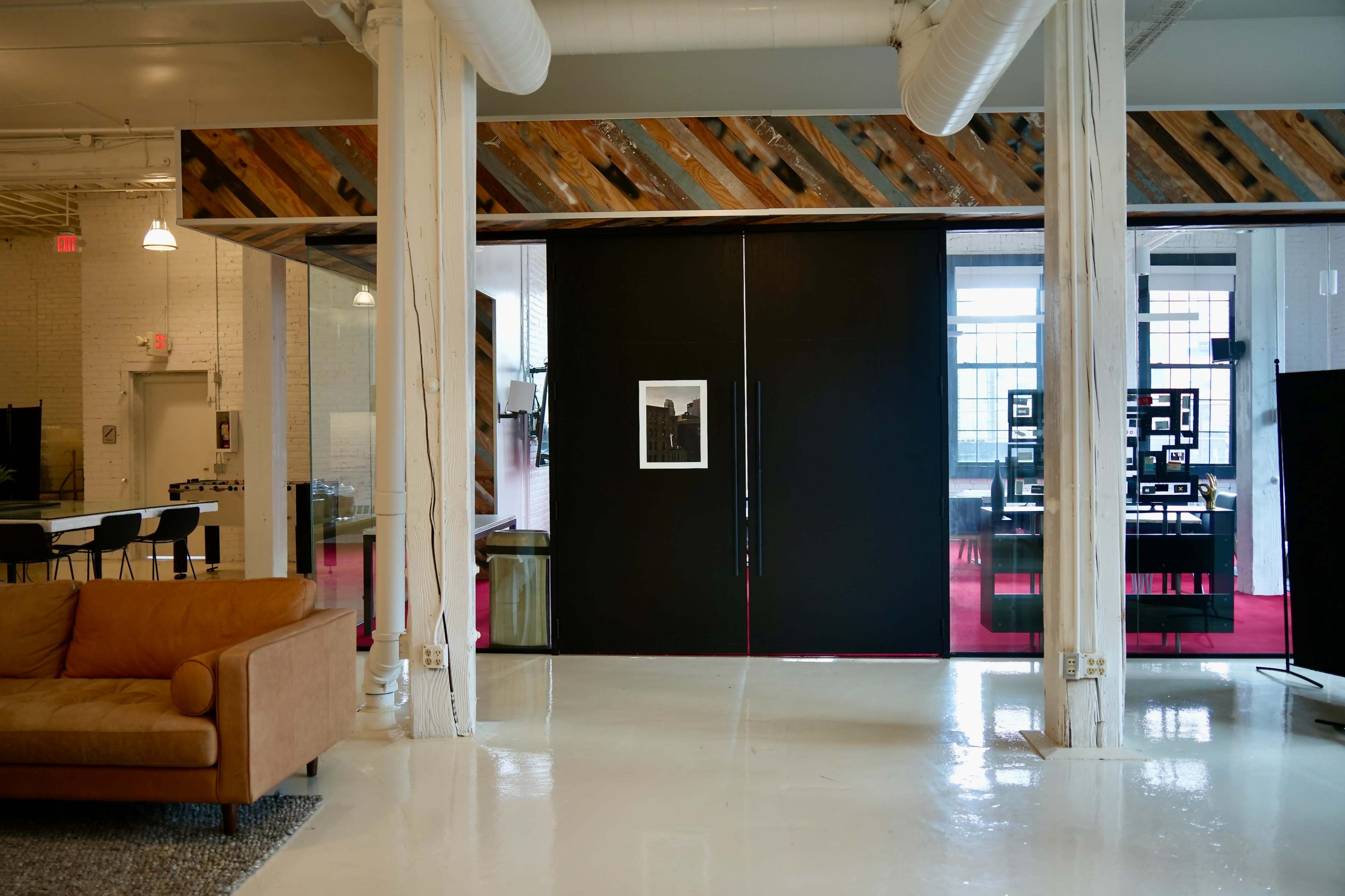 The image shows a modern interior space with a large black sliding door, a cozy tan sofa in the foreground, and a bright open area that includes office furnishings in the background.