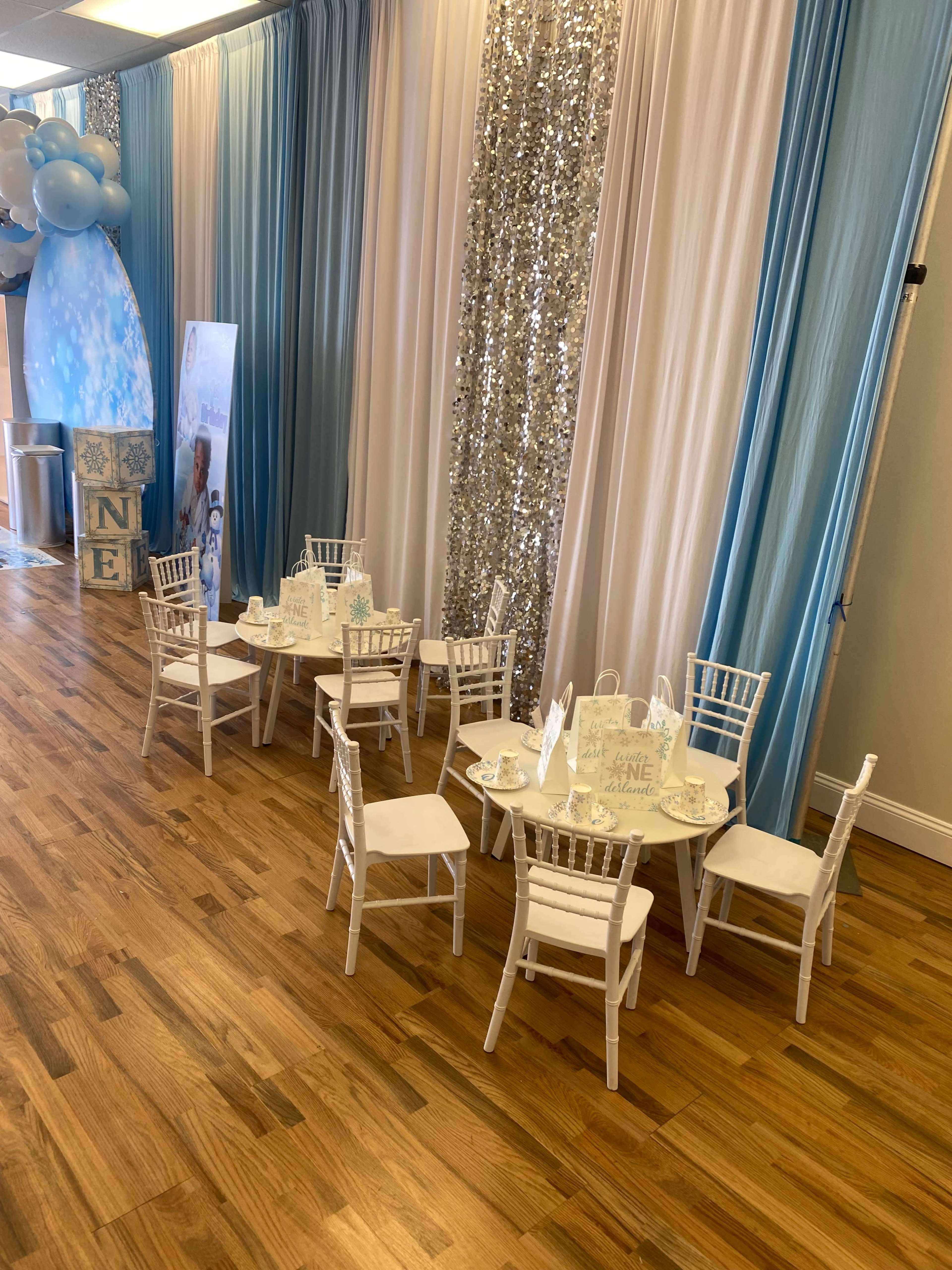 A small dining setup featuring white chairs and tables arranged against a backdrop of blue and silver drapes in a well-lit event space.