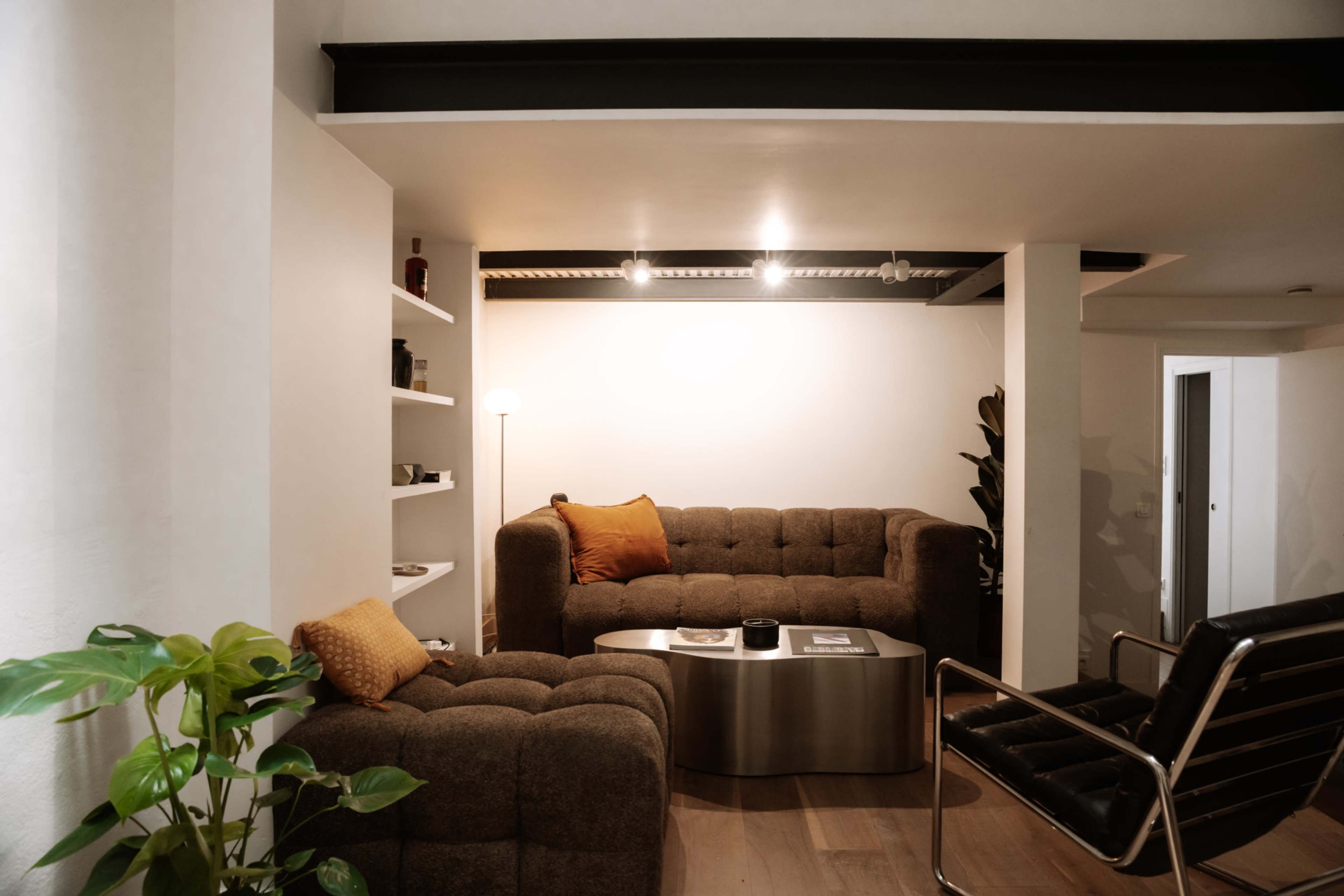 A cozy living room features two brown couches, a round metal coffee table, and a floor lamp, with a plant in the foreground and shelves displaying various items on the wall.