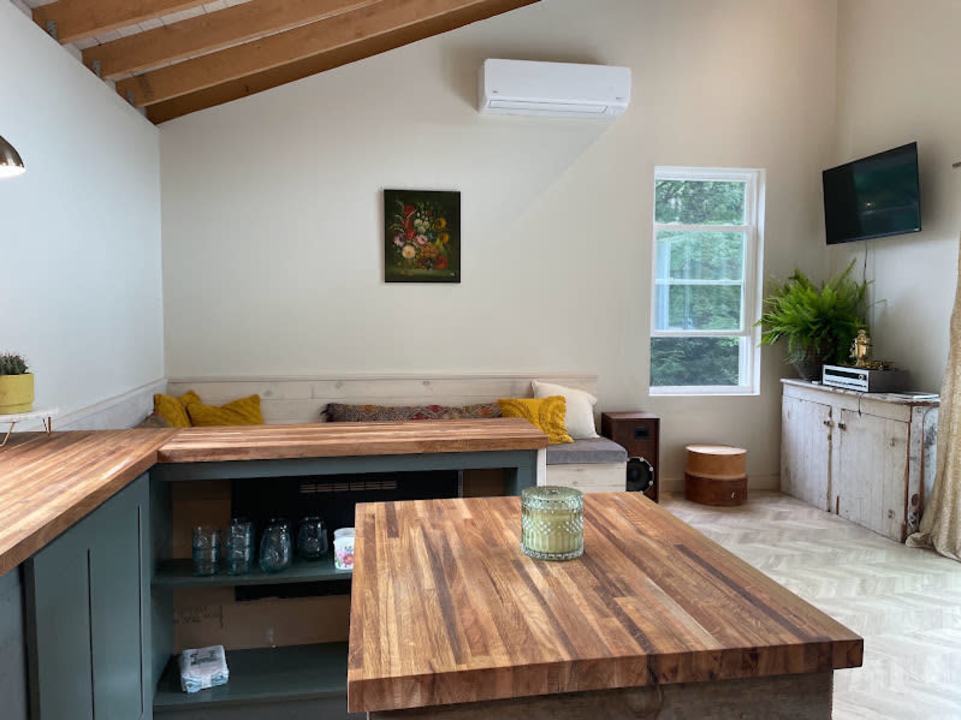 The image shows a cozy living area with a wooden table, a seating nook with cushions, and a TV mounted on the wall, surrounded by natural light and greenery.