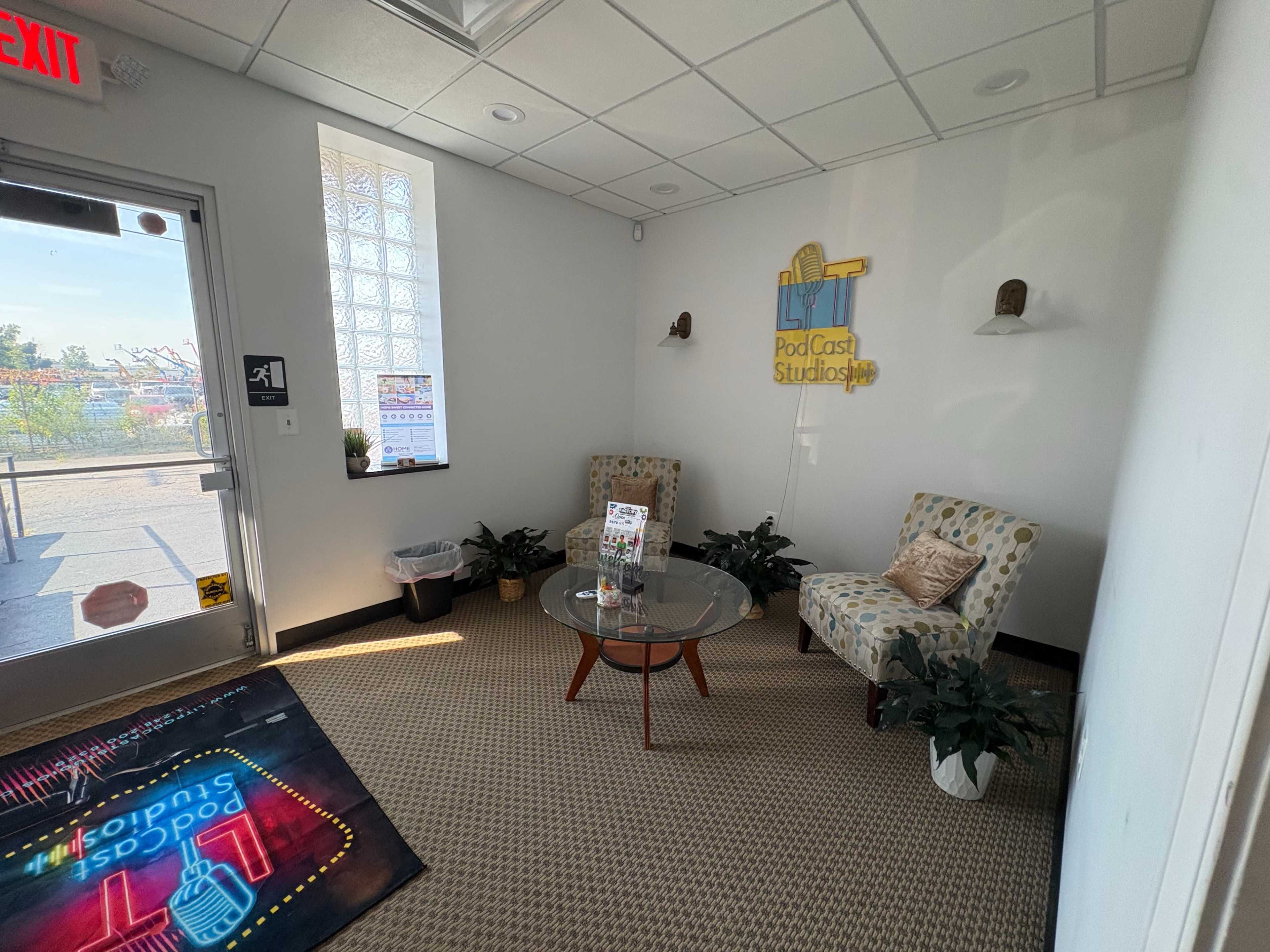 The image shows a waiting area in a studio, featuring two chairs, a small table with a brochure, and plants, with a sign on the wall that reads "Podcast Studios."