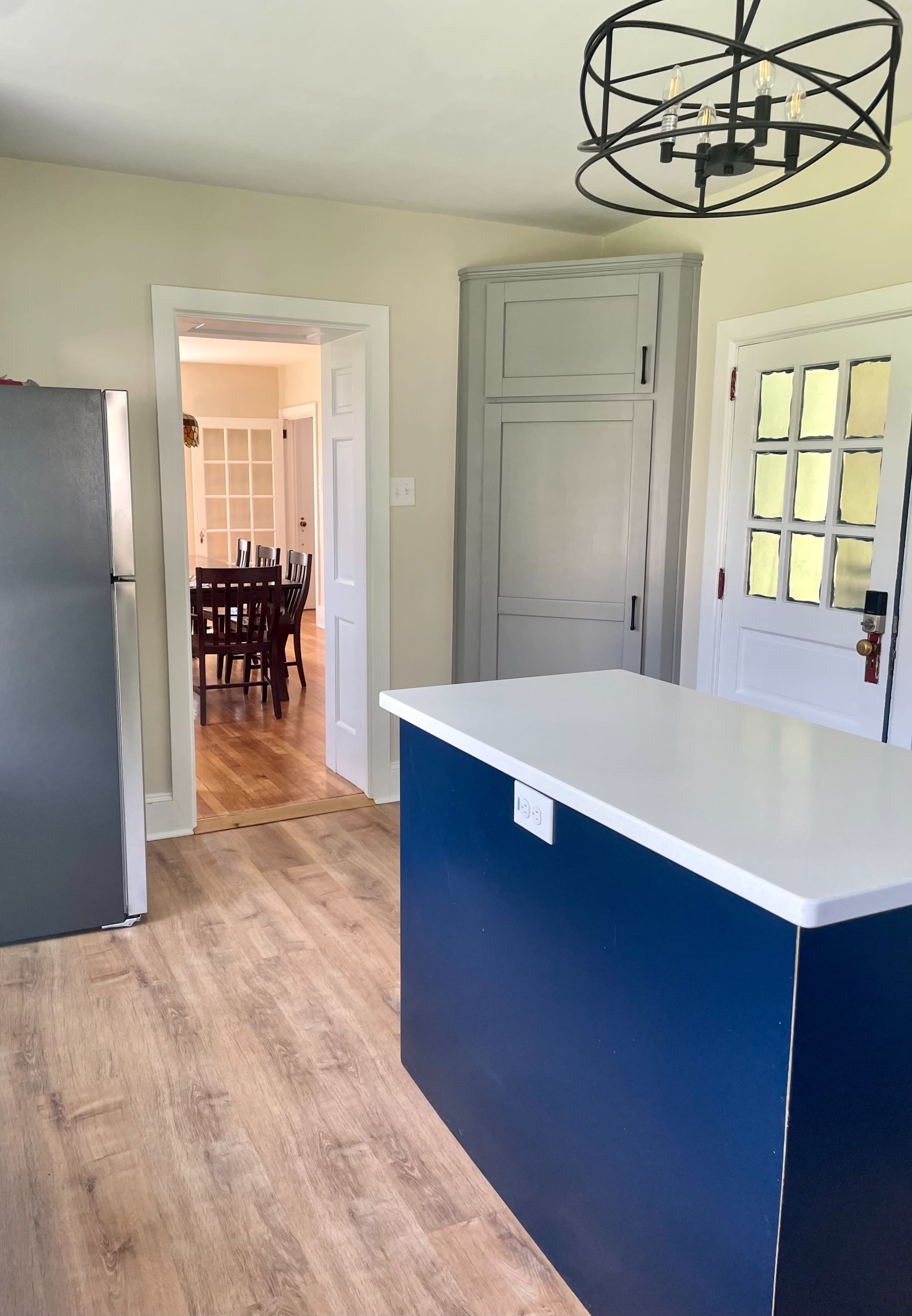 A modern kitchen with a blue island, stainless steel refrigerator, and an entrance to a dining area in the background.