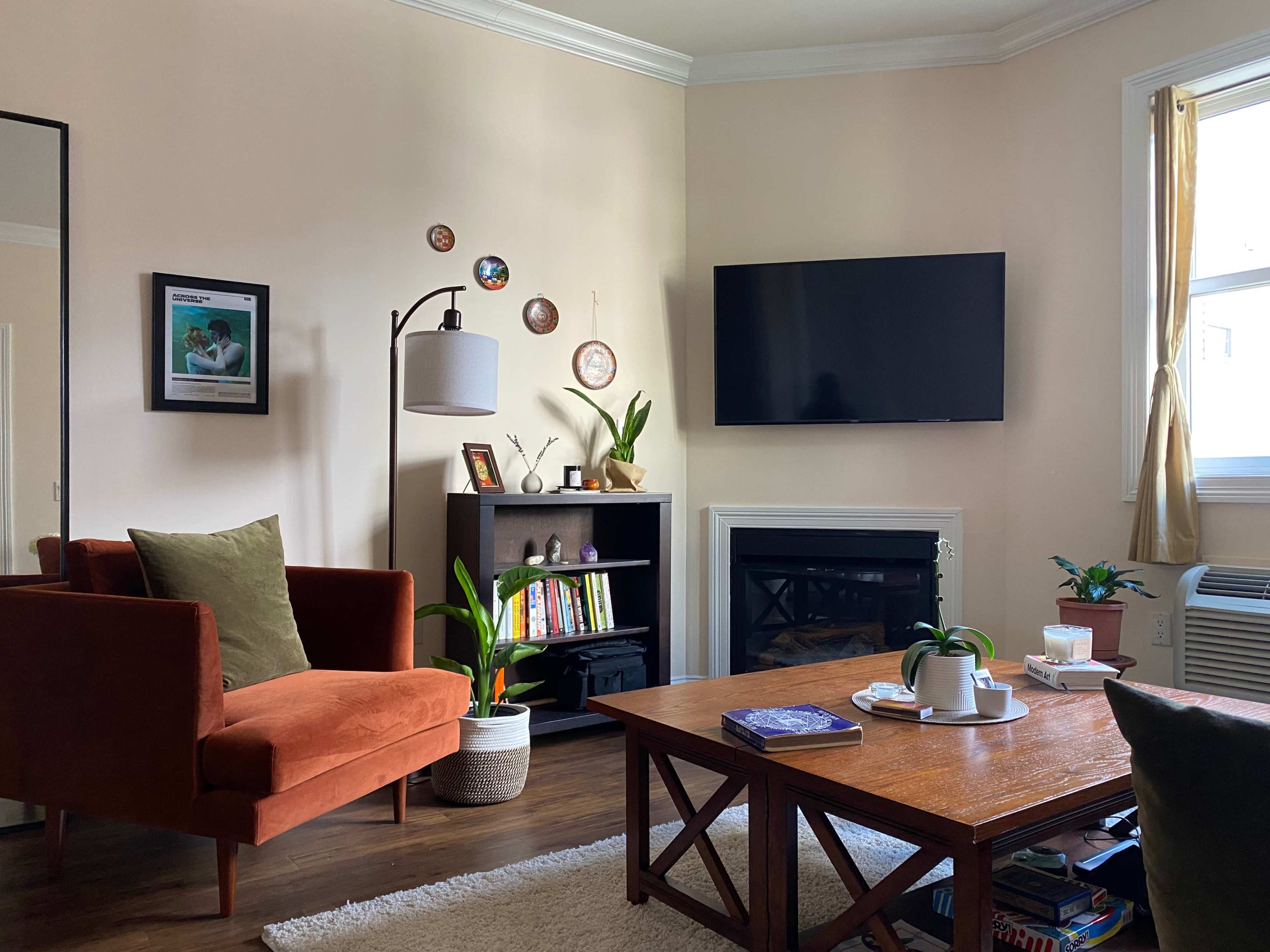 The living room features an orange armchair, a wooden coffee table, a bookshelf, and a mounted television above a fireplace.