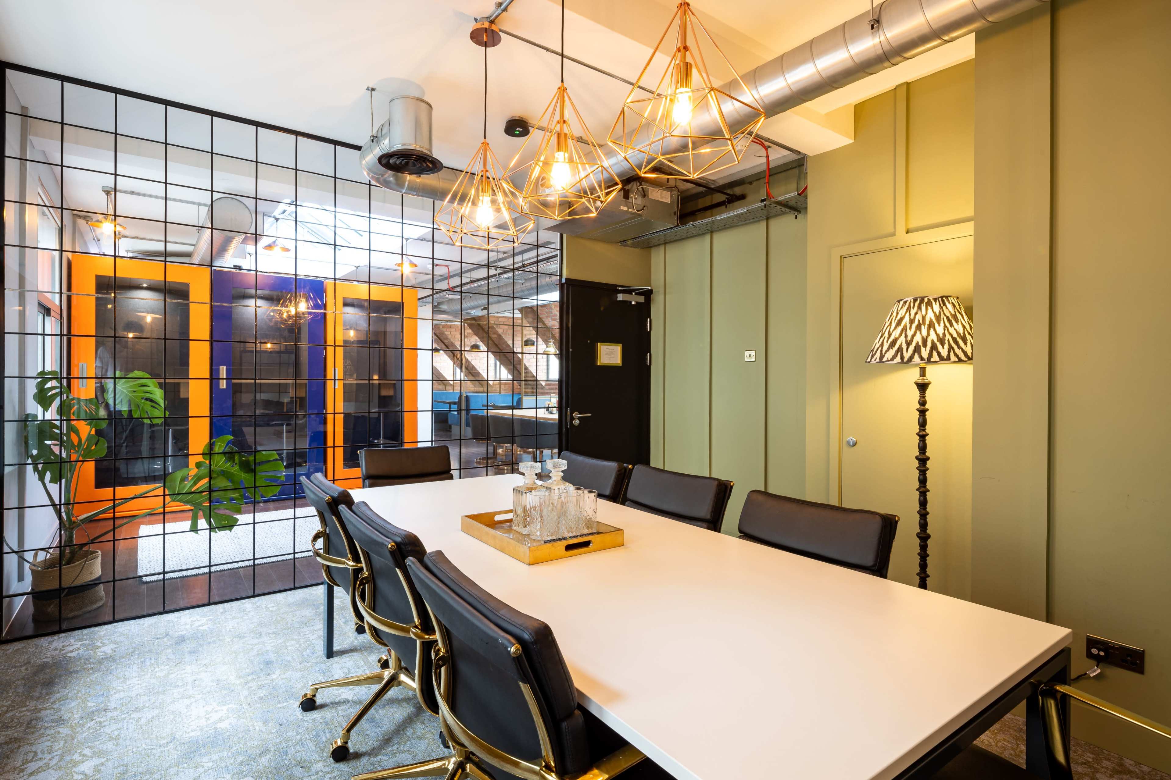 A modern conference room features a large white table surrounded by black chairs, with geometric pendant lights and a glass partition leading to a space with orange doors.