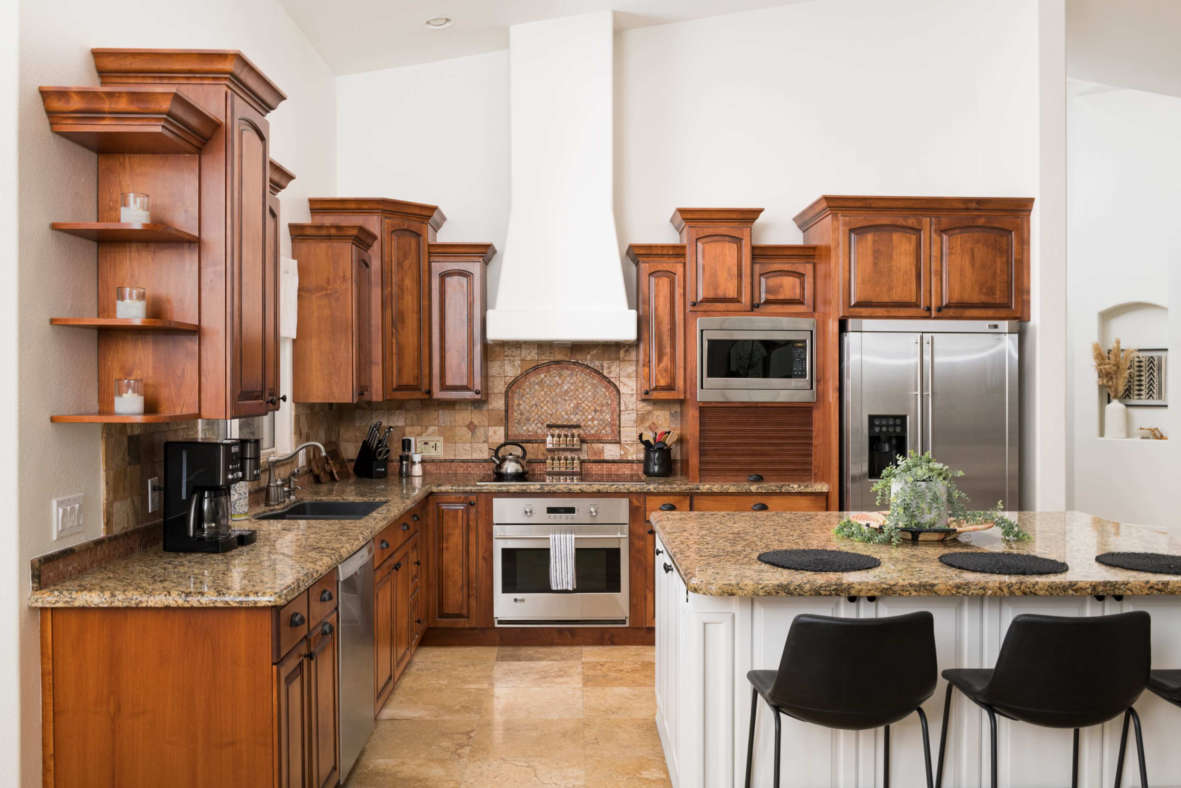 A modern kitchen features wooden cabinetry, granite countertops, and stainless steel appliances, with a center island and bar stools.