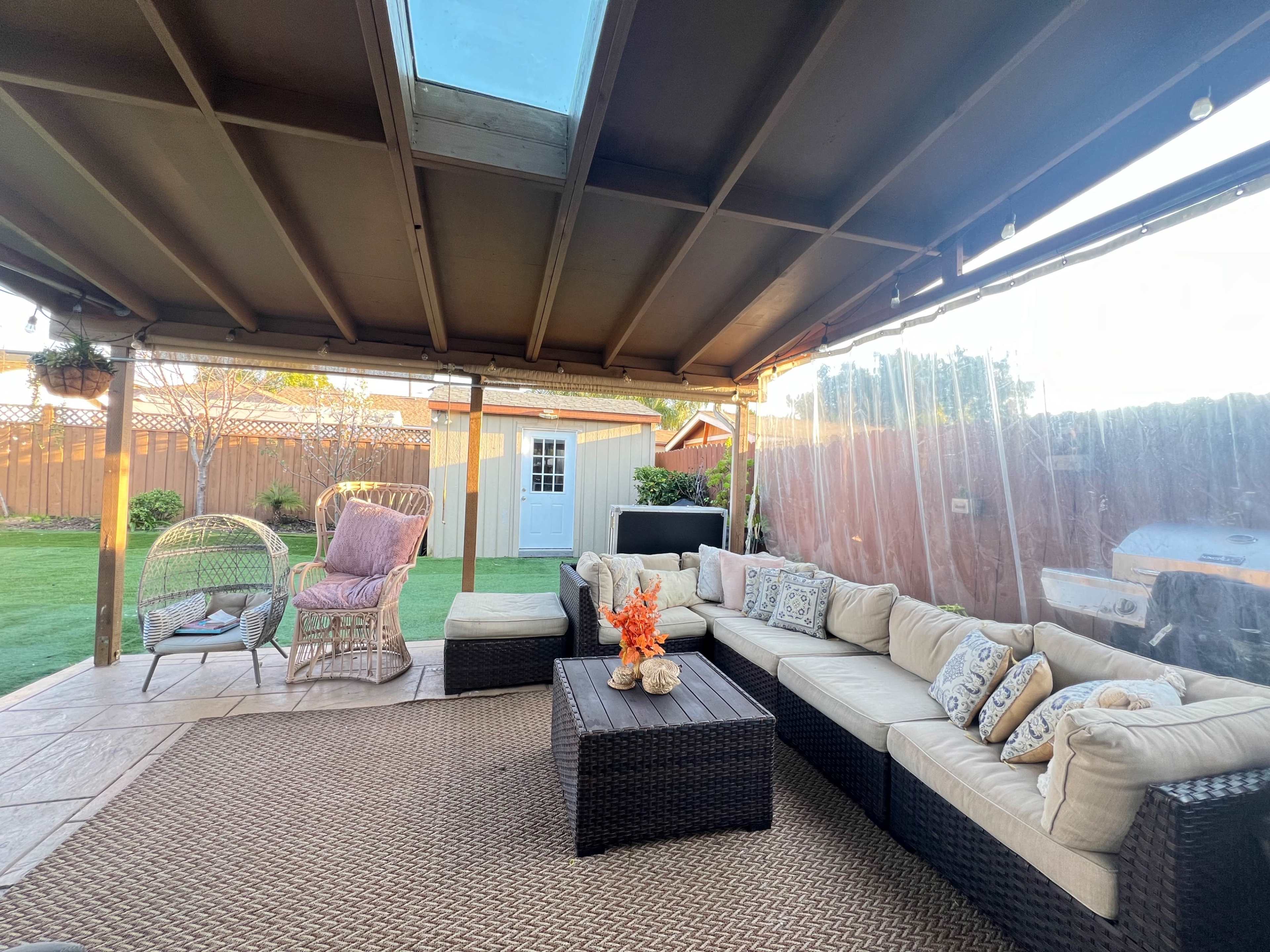 A covered outdoor seating area with a sectional sofa, a coffee table, and decorative elements, adjacent to a grassy backyard with a shed in the background.