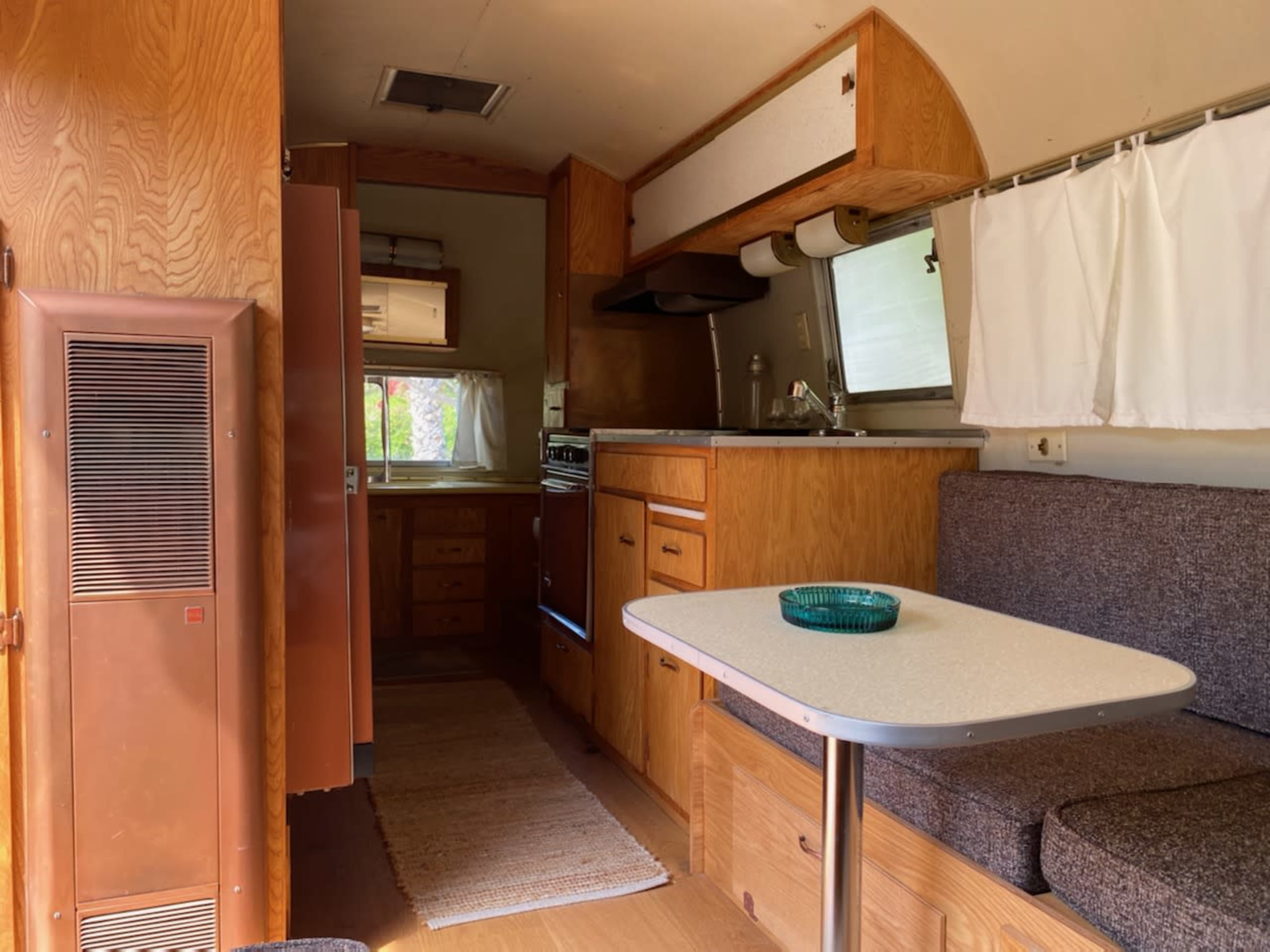 The interior of a retro camper features wooden cabinets, a kitchenette, a dining area with a table, and a soft seating area.