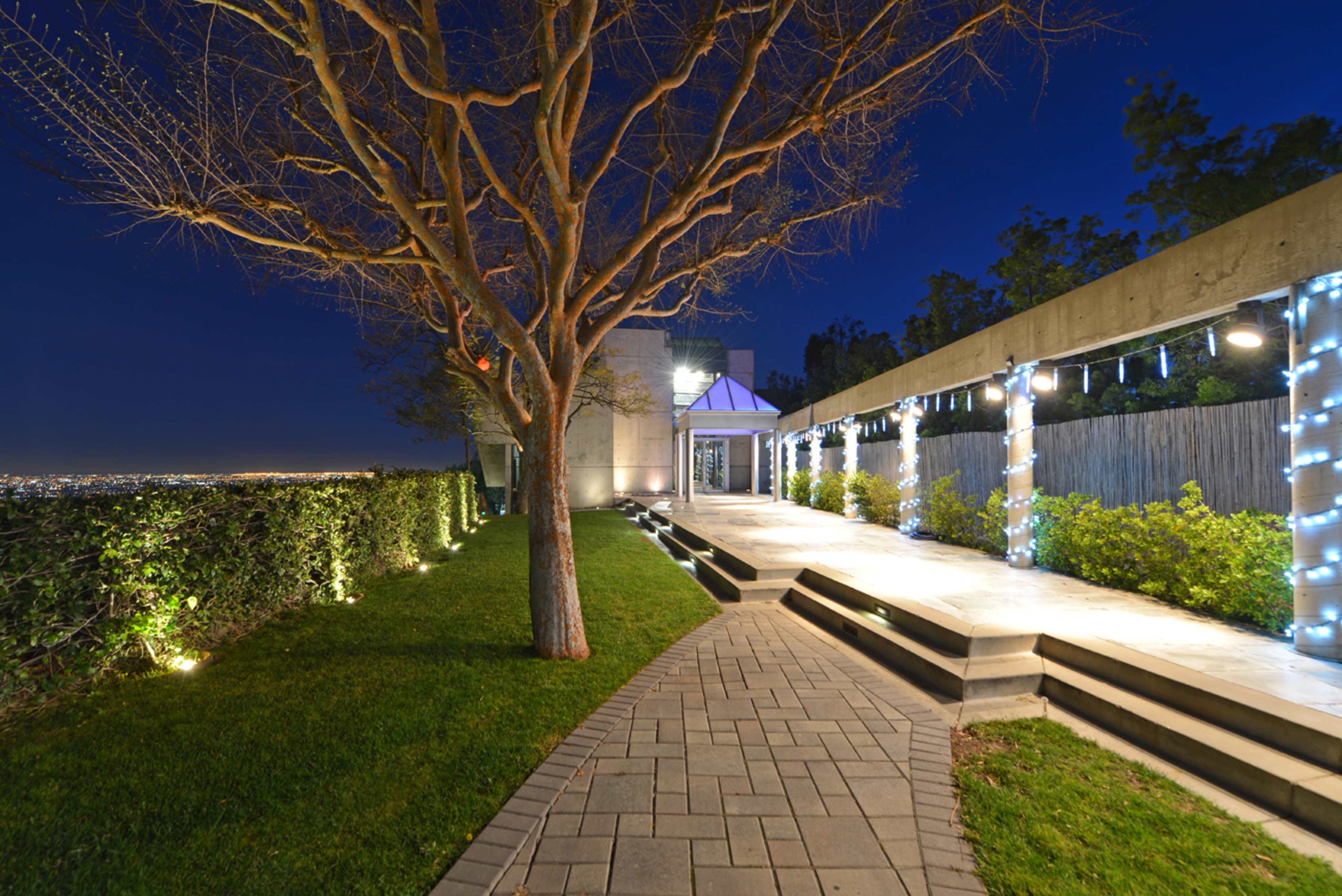 A winding stone path leads from a bare tree to a modern building, illuminated by lights strung along a covered patio, with city lights visible in the background.