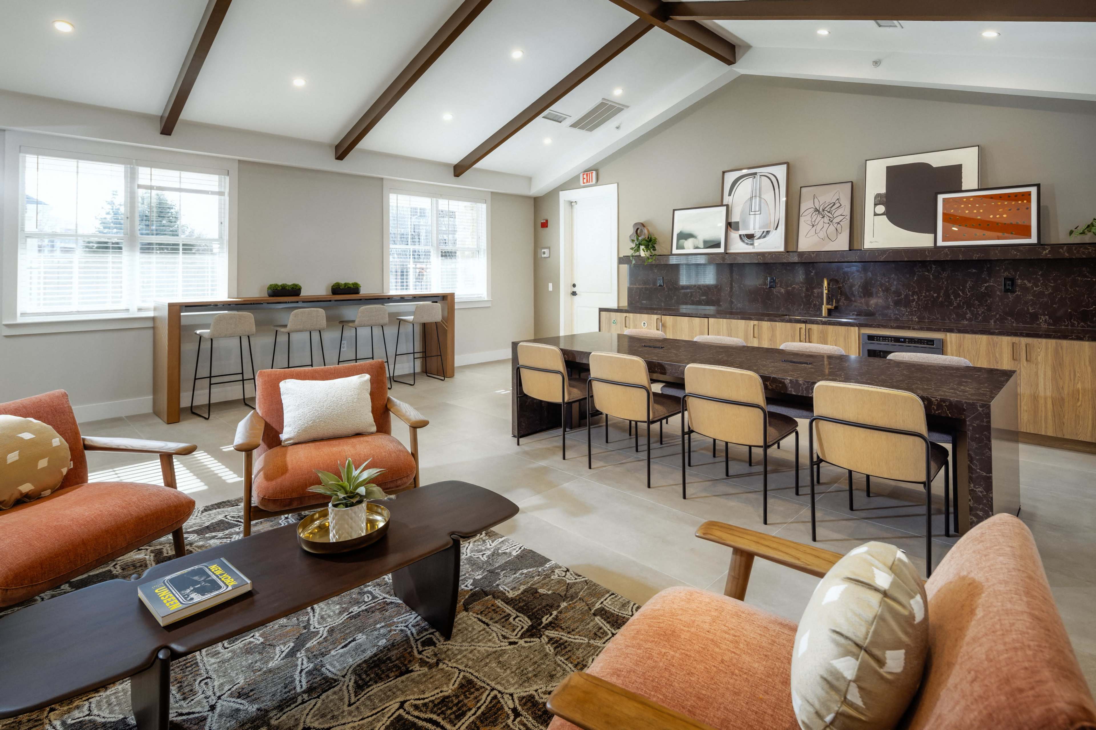 A modern communal space features a dining area with a long marble table, upholstered chairs, and a cozy sitting area with orange sofas and a patterned rug.