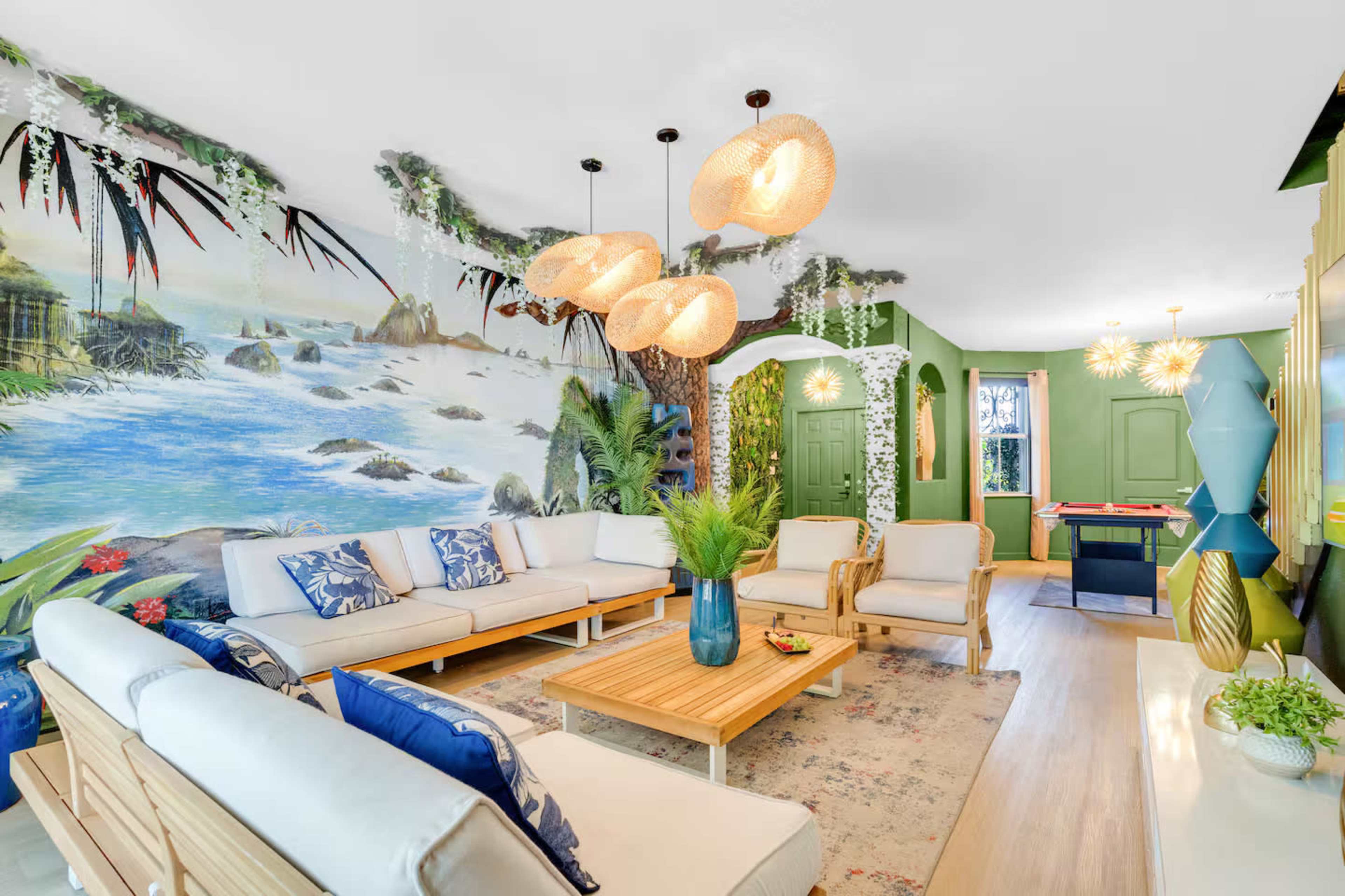 The living room features a mural of a coastal scene, modern furniture, and pendant lights, with a game area visible in the background.