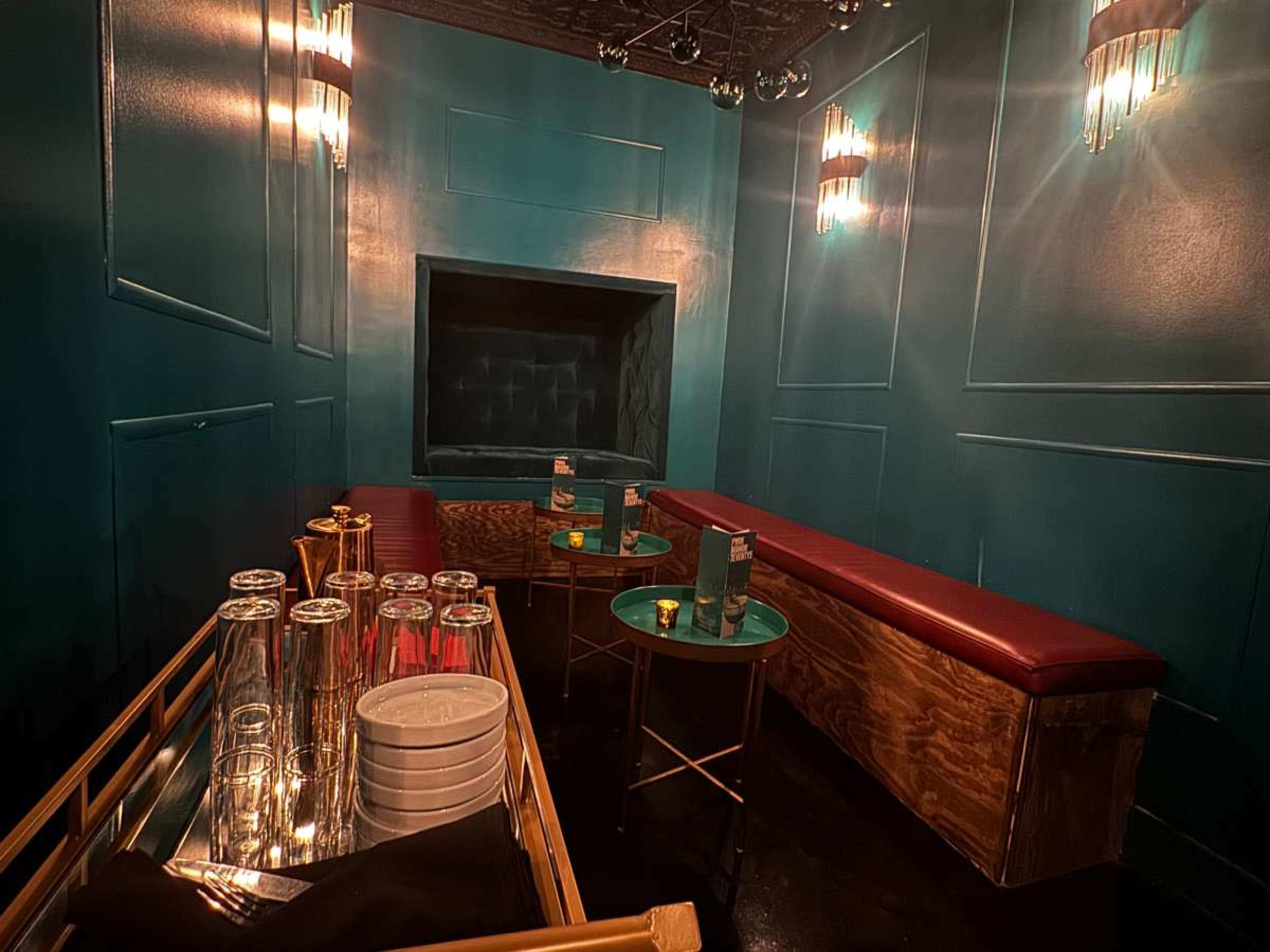The image shows a small, dimly lit lounge area with teal walls, red seating, and a bar cart holding various glassware.