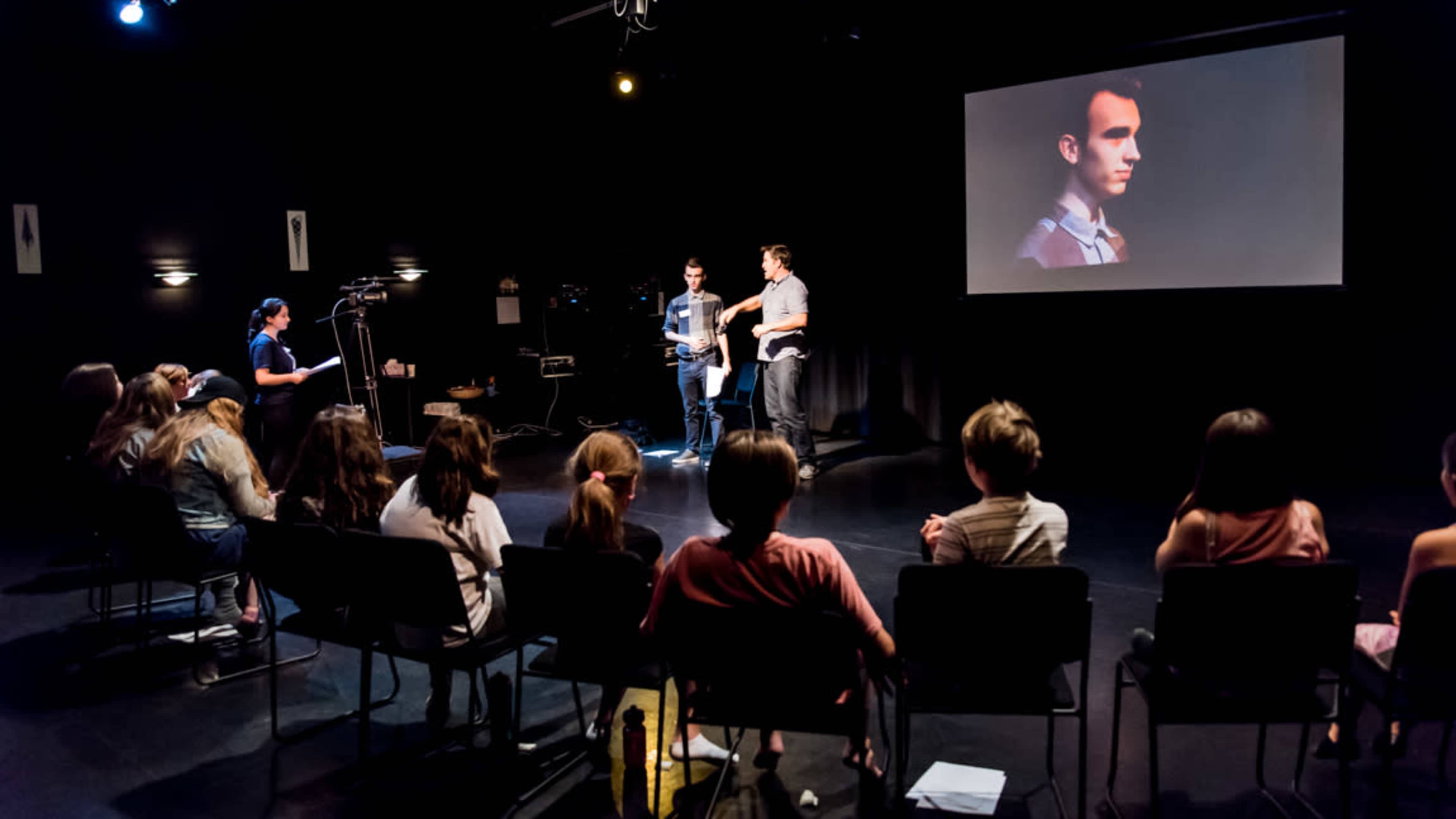A small group of students watches a presentation in a darkened room with a large screen displaying a man's profile.