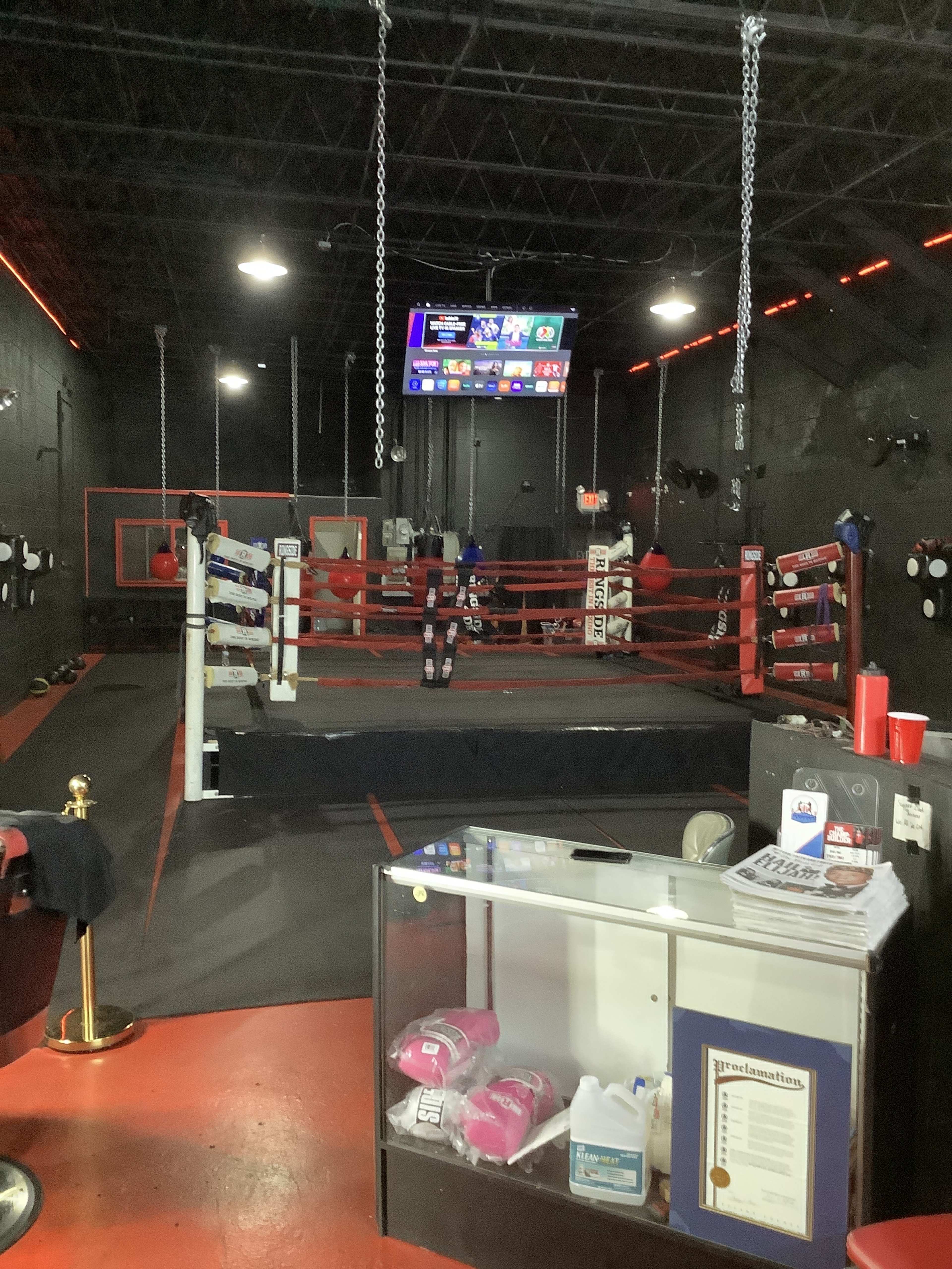 A boxing gym interior with a boxing ring in the center, illuminated by red and white lights, and a reception area with a glass display case in the foreground.