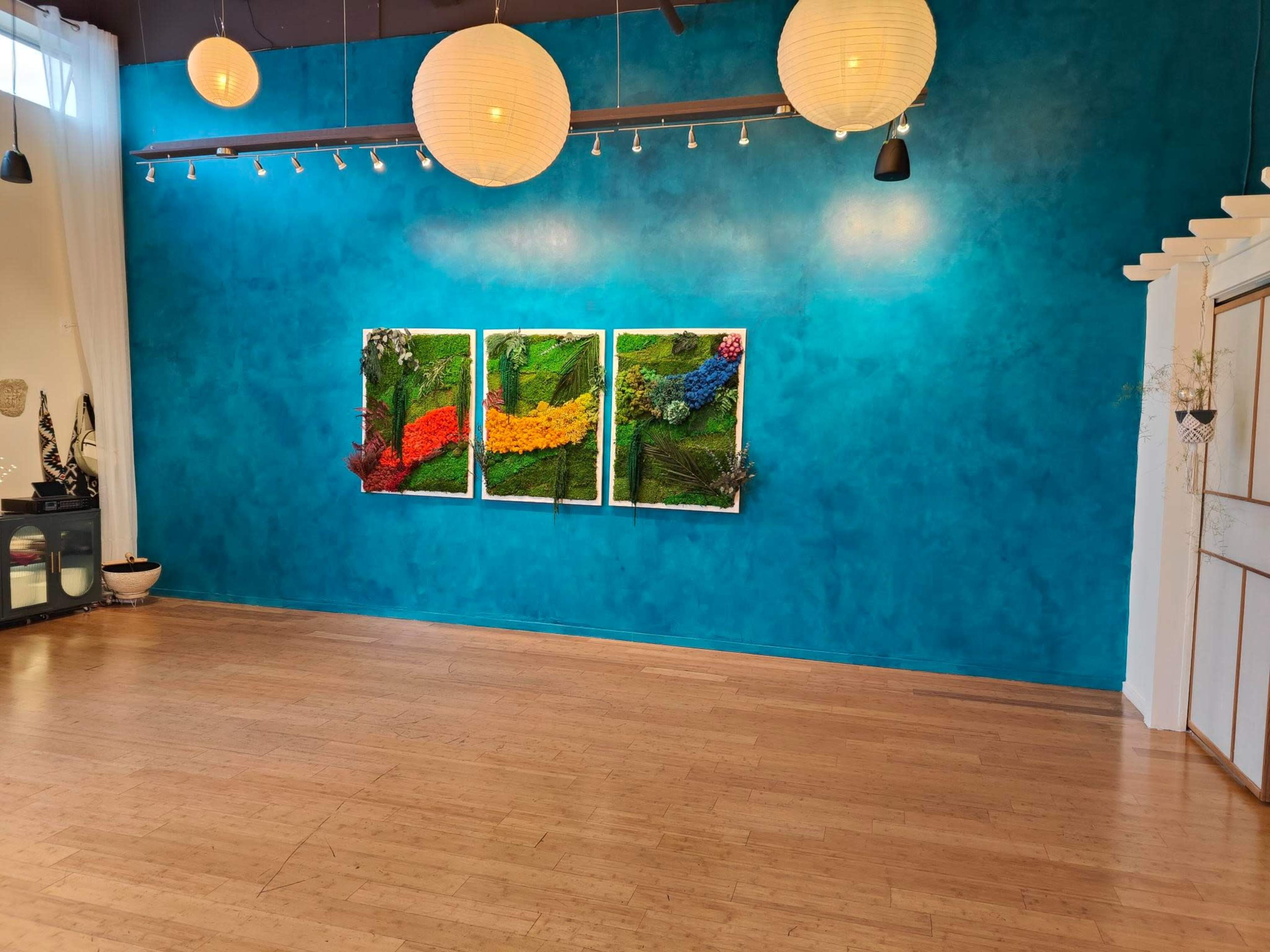 A spacious room with a bright blue wall featuring three framed artworks made of colorful flowers and plants, illuminated by hanging paper lanterns.