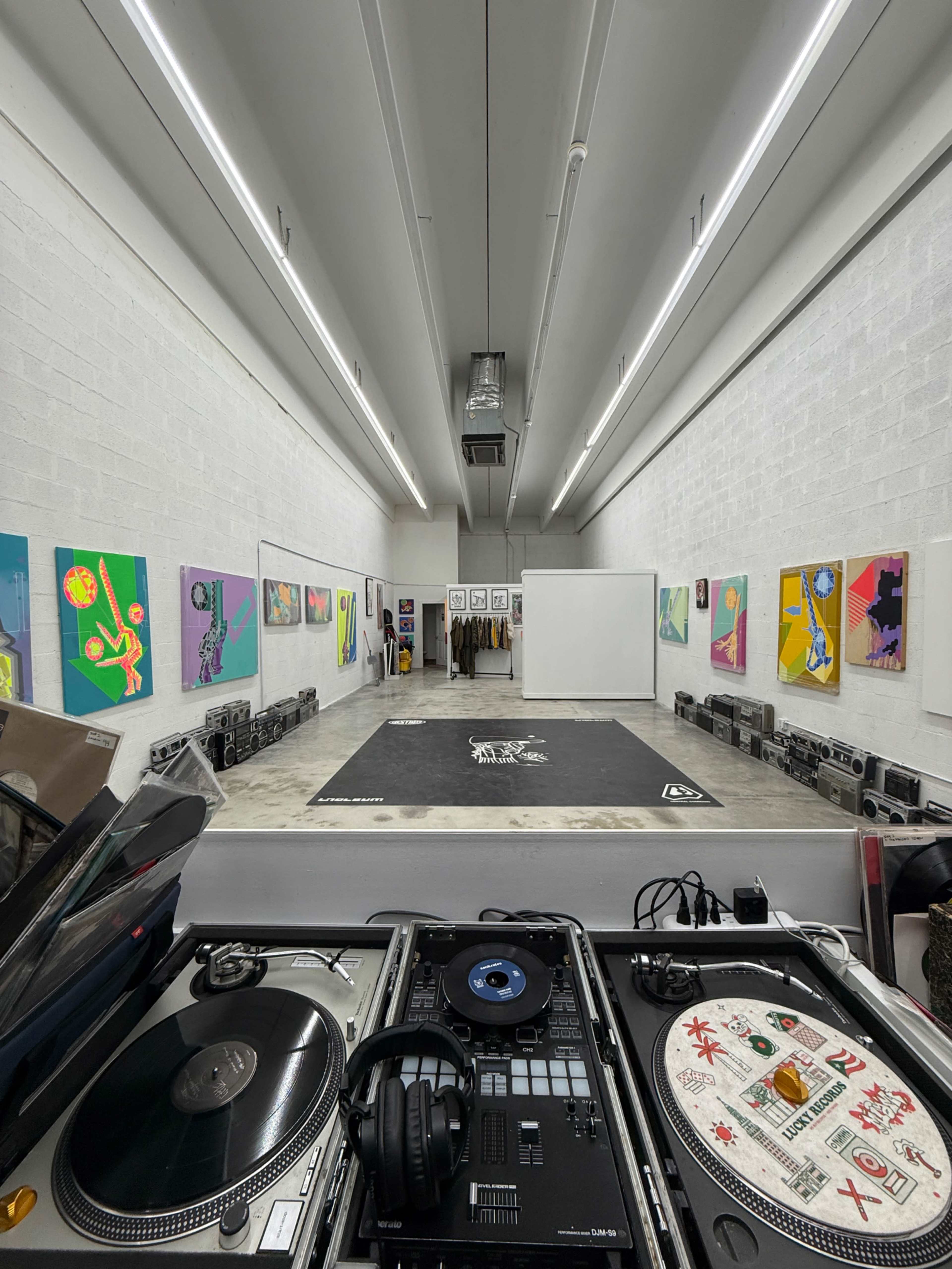 The image shows a long, narrow art gallery with vibrant artworks displayed on the walls and a DJ setup in the foreground.