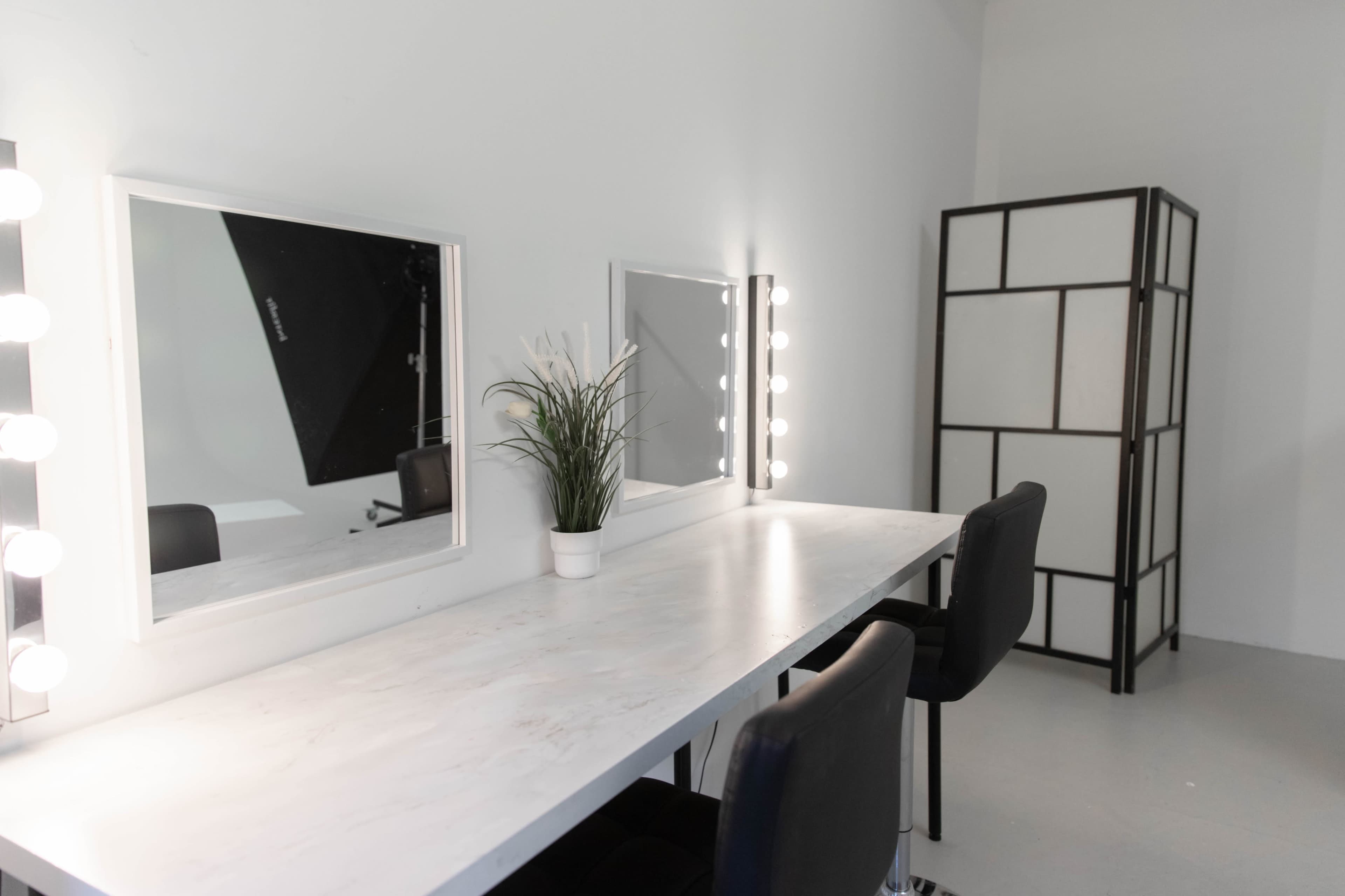 The image shows a minimalist dressing room with a long white table, two mirrors with bulbs, and a potted plant, along with a black folding screen in the background.