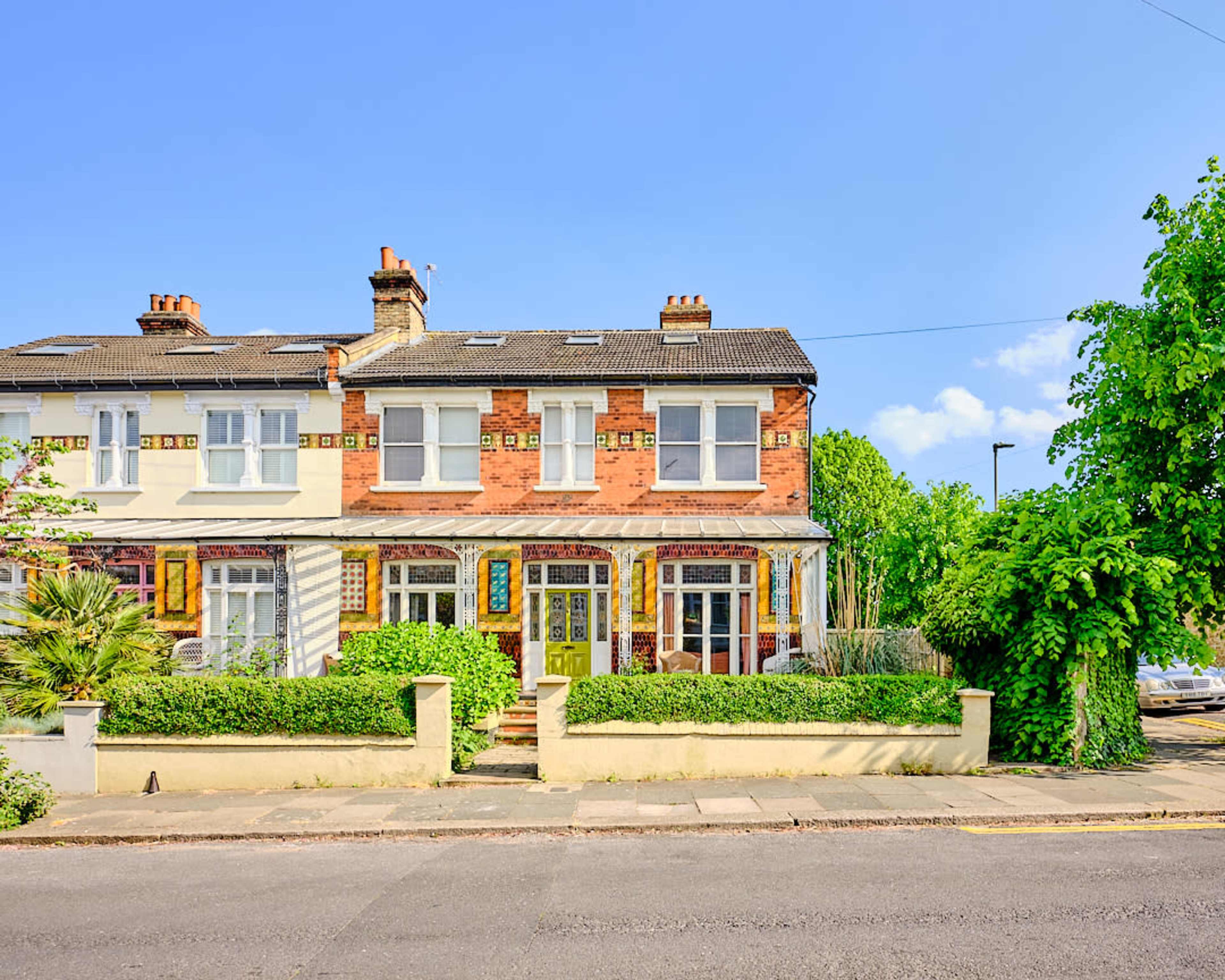 The image shows a two-story, brick-covered end-of-terrace house with a colorful facade and a neatly trimmed garden.