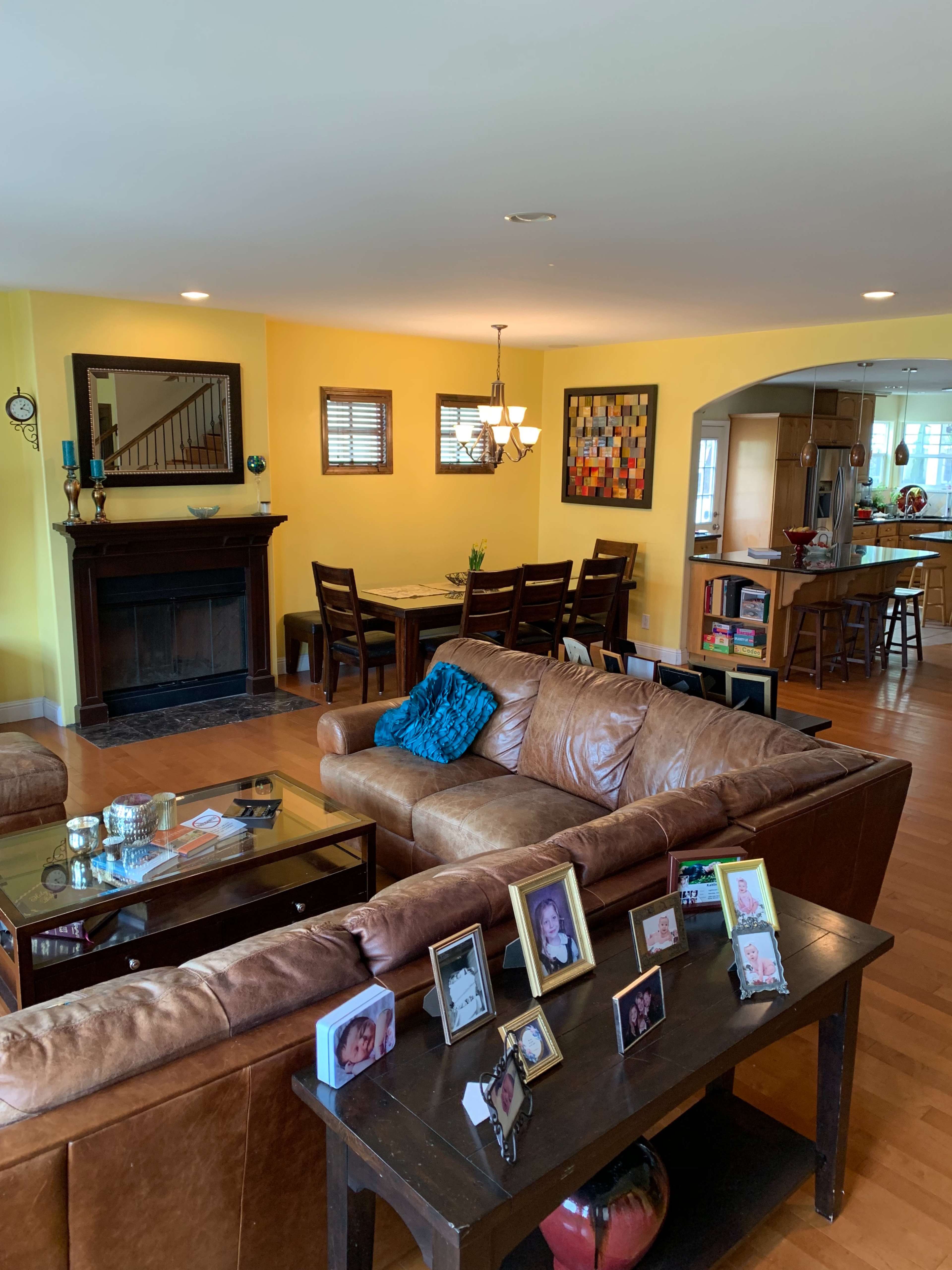 The image shows a living room with a brown leather couch, a coffee table, and a dining area in the background, featuring framed photographs on a side table.