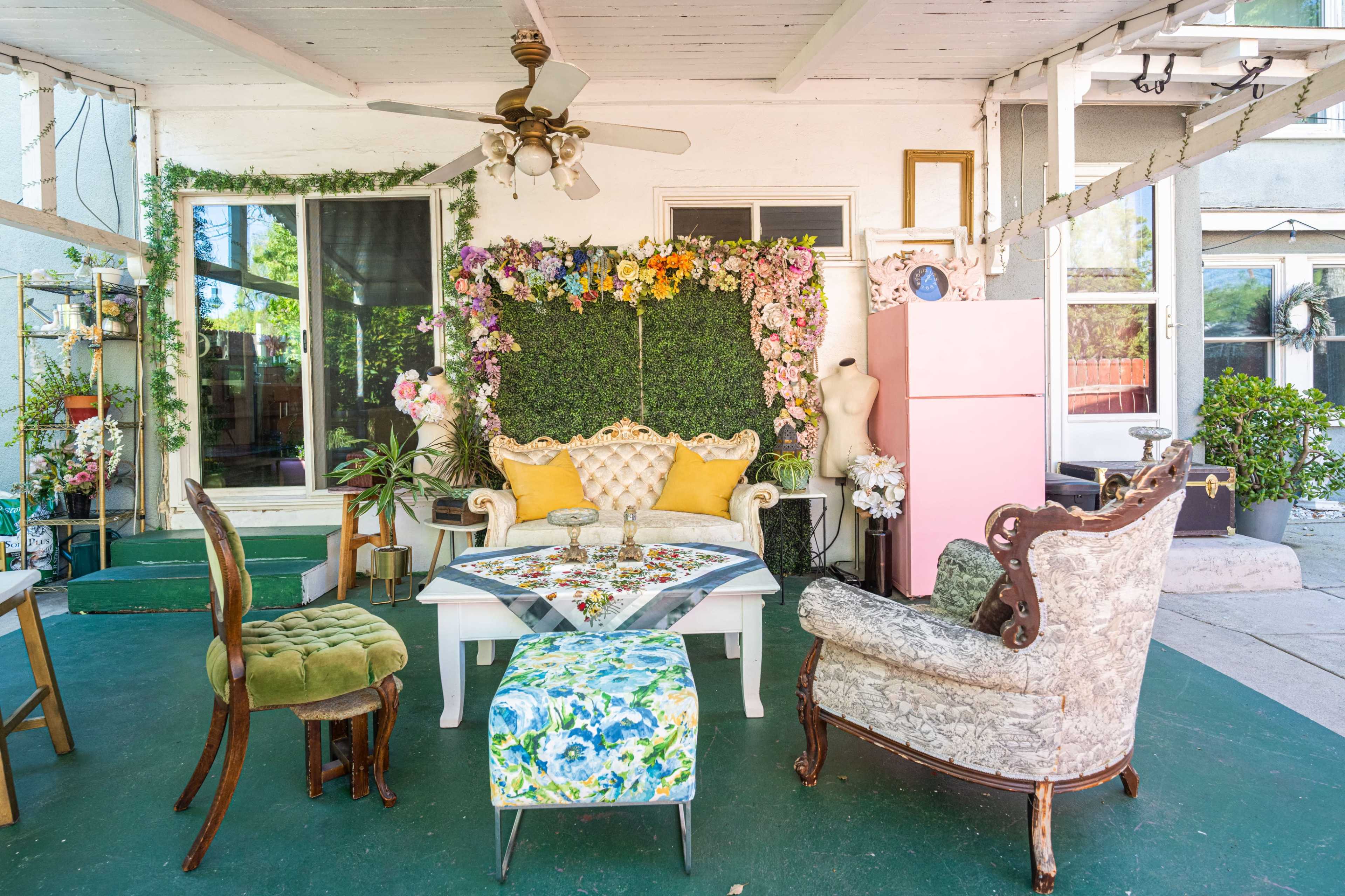 The image shows a decorated outdoor patio with a sofa, two chairs, a set table, and a backdrop of flowers and greenery.