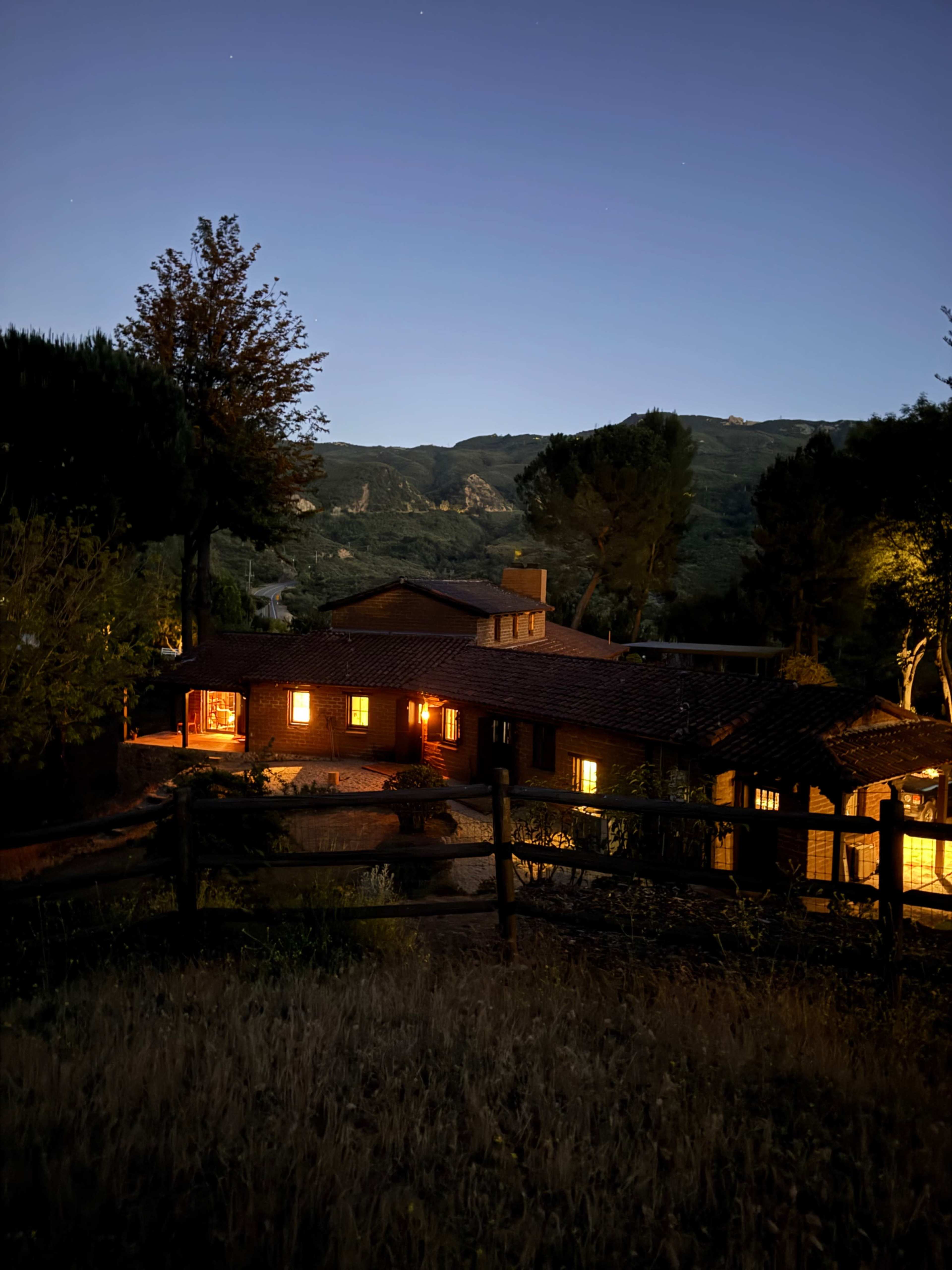A brick house is surrounded by trees and mountains, with warm lights glowing from its windows at twilight.