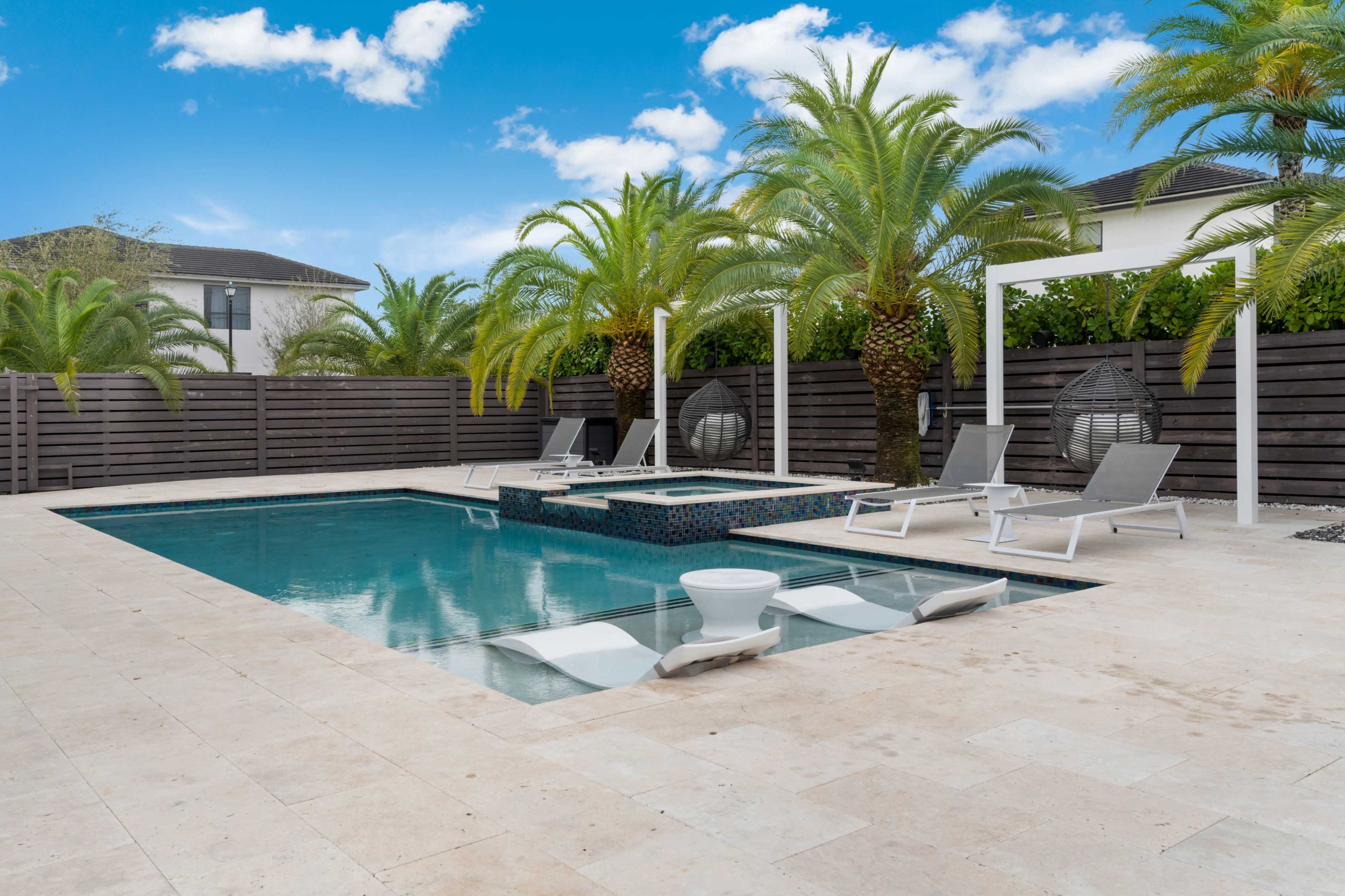 A backyard area featuring a rectangular swimming pool with a spa, surrounded by lounge chairs and palm trees against a clear blue sky.