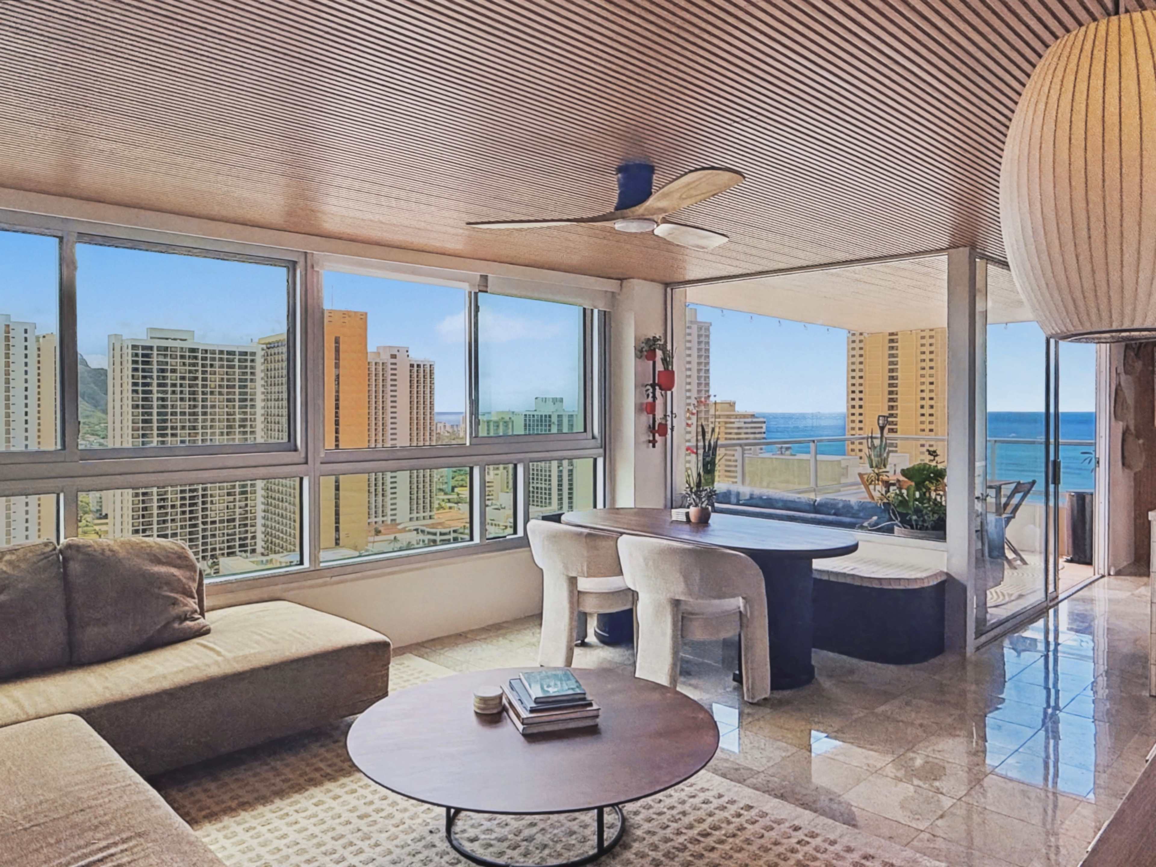 A modern living space features large windows with a view of a city skyline and ocean, furnished with a beige sofa, round coffee table, dining area, and ceiling fan.
