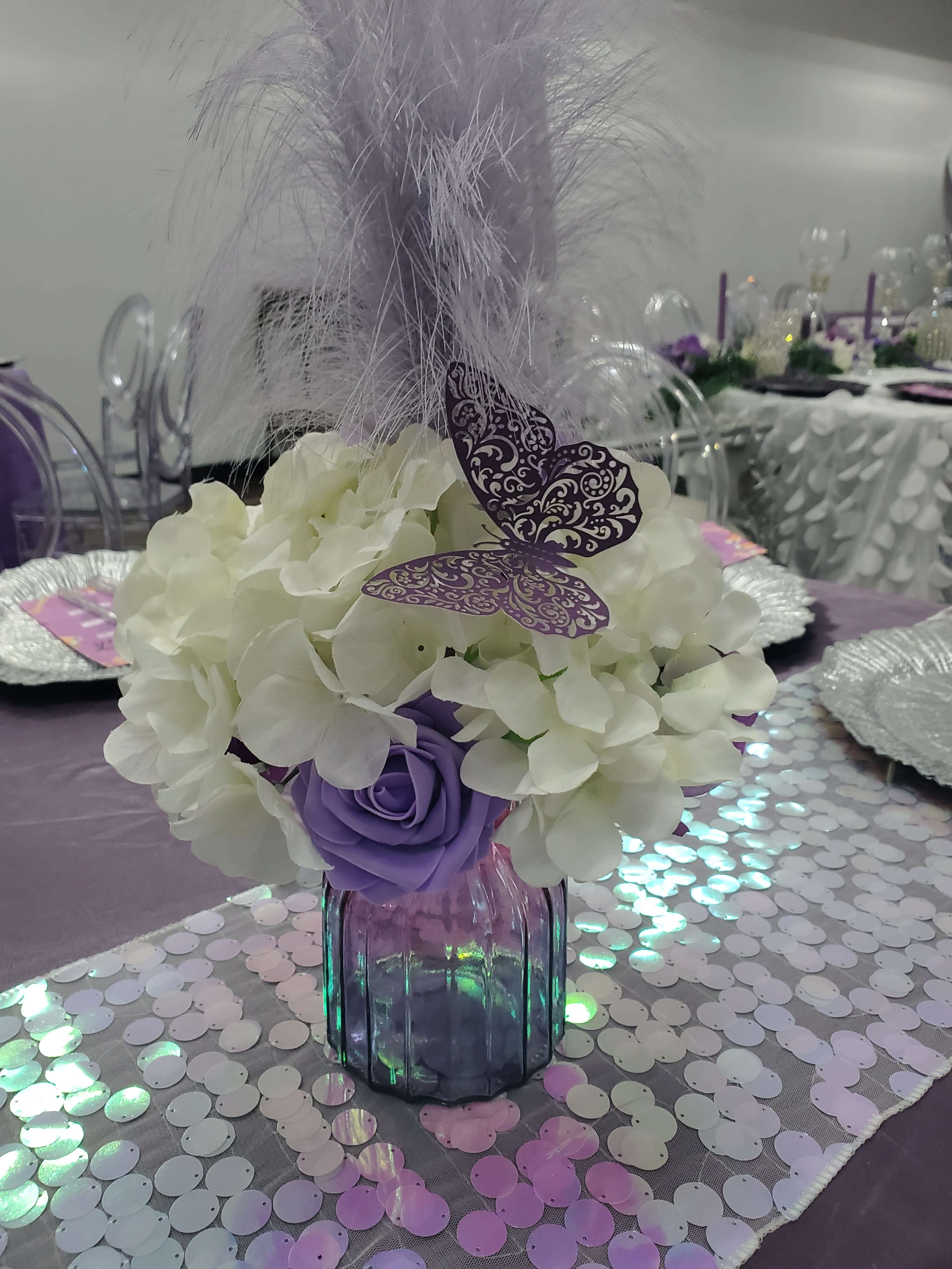 A floral centerpiece featuring white hydrangeas, a purple rose, and a decorative butterfly, placed in a glass jar on a shimmering table runner.